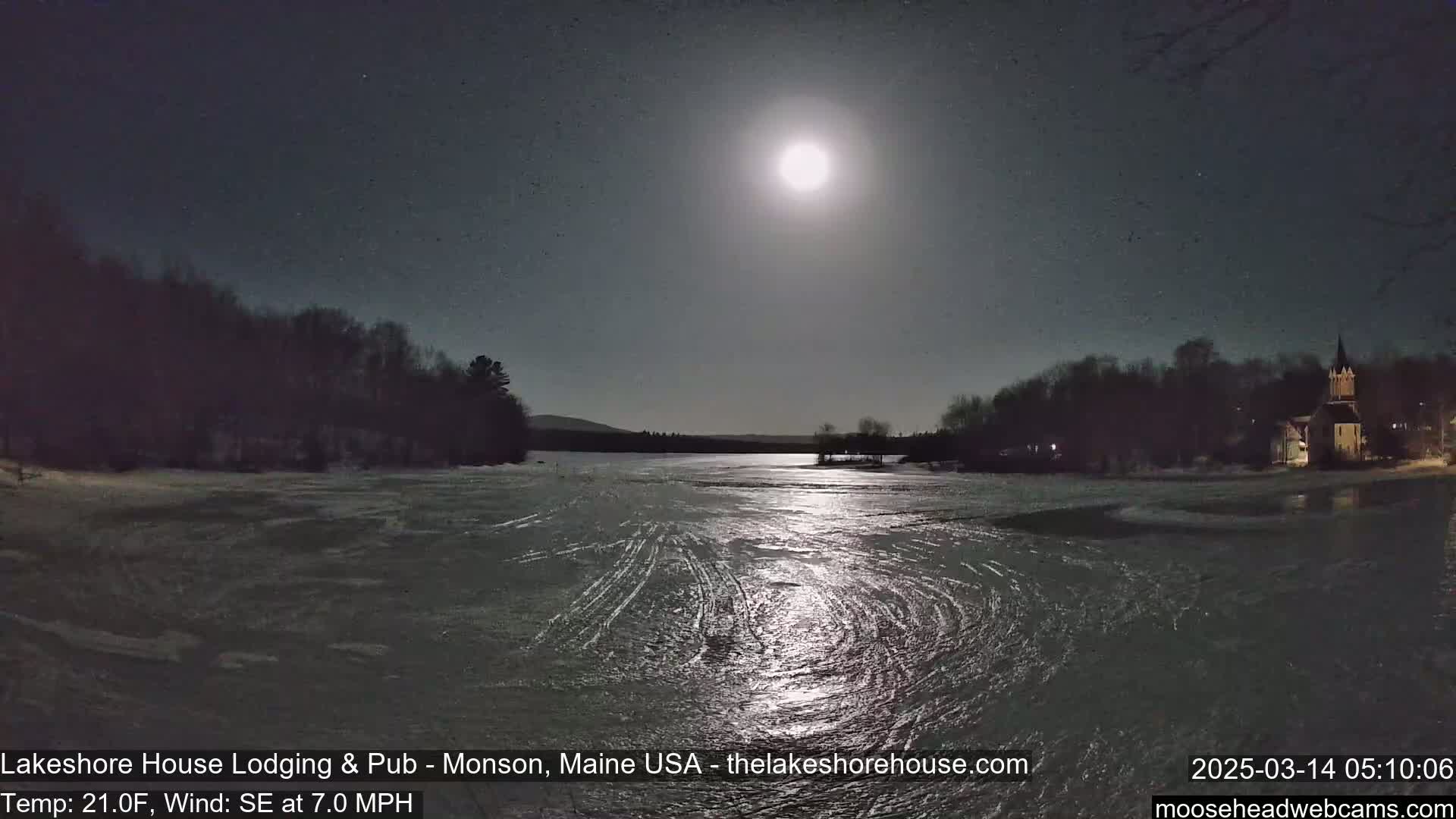 A bright moon illuminates a frozen lake under a clear night sky, with trees and a church visible on the far shore.
