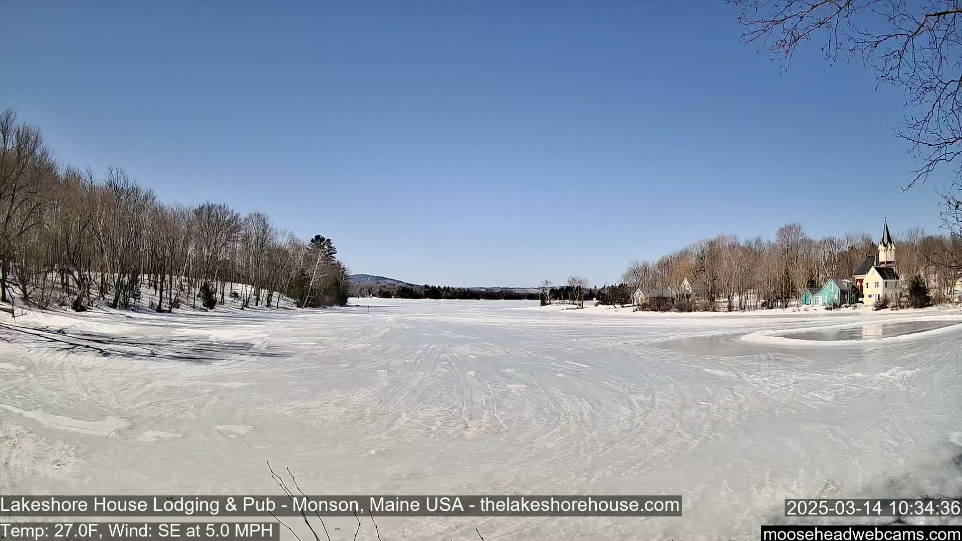 A frozen lake, with a village and bare trees on the far shore, under a clear blue sky.