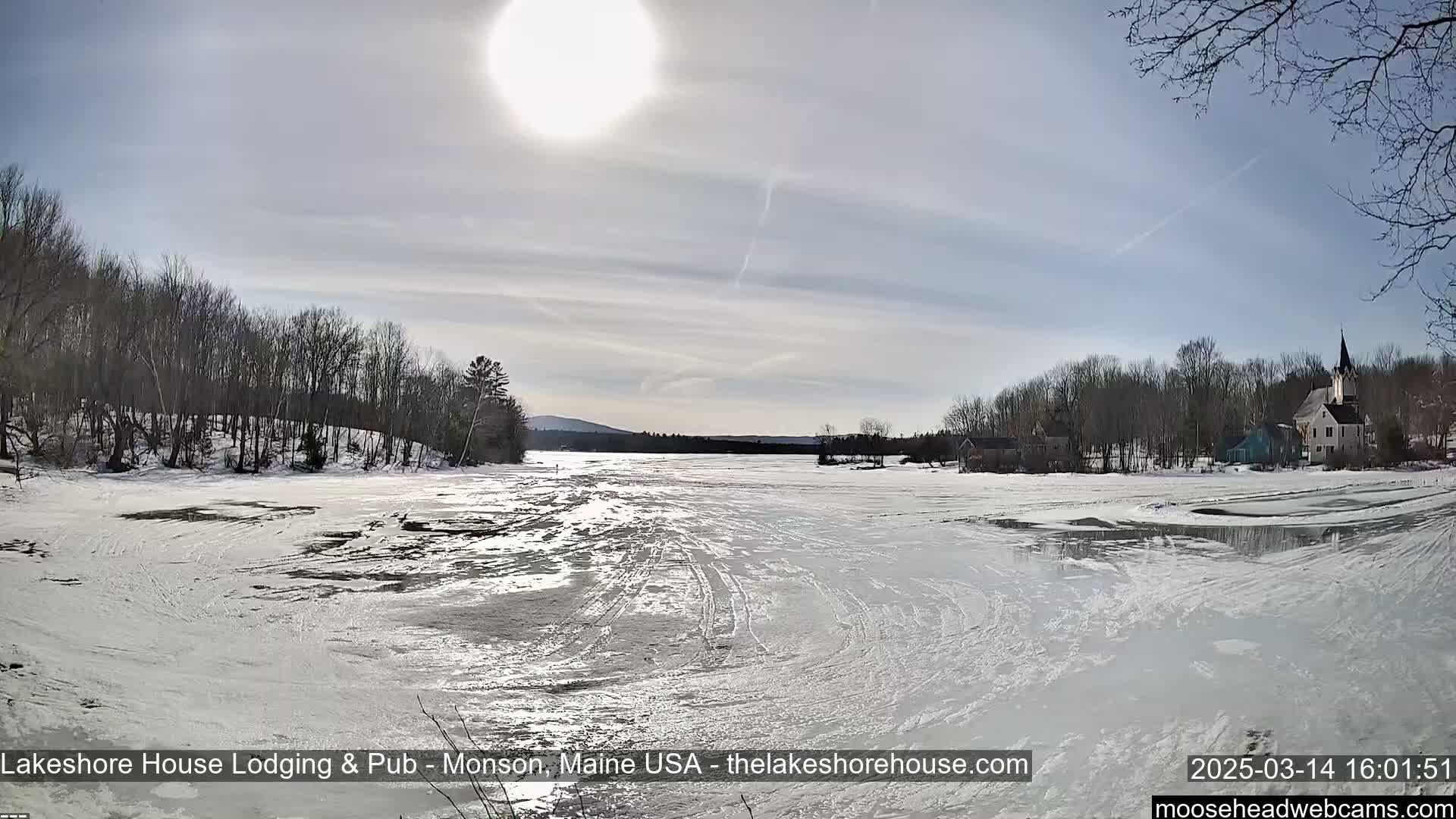 A mostly sunny day reveals a frozen lake with tire tracks, snow-covered banks lined with bare trees, and a small village in the distance.