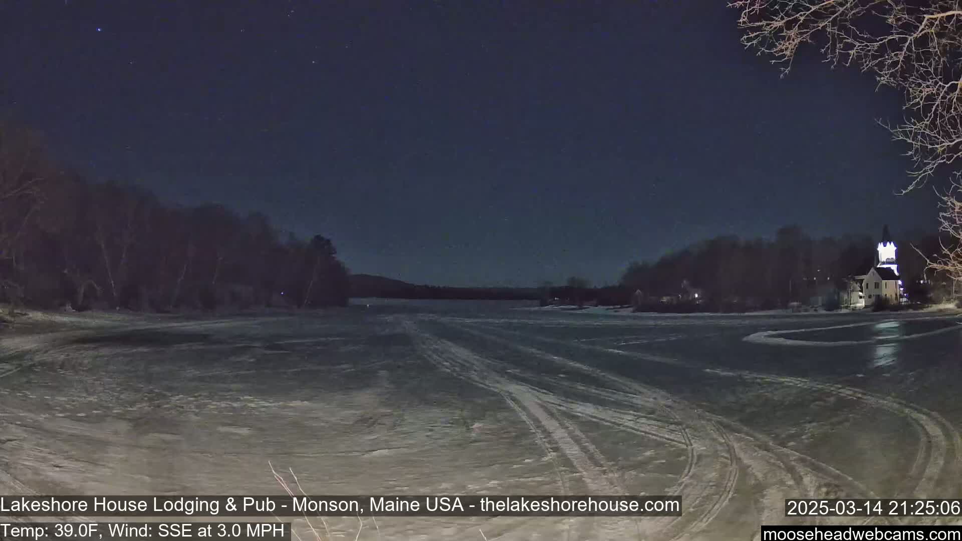 A frozen lake at night under a starry sky shows tire tracks across its surface, with a shoreline of dark trees and a building lit up in the distance.