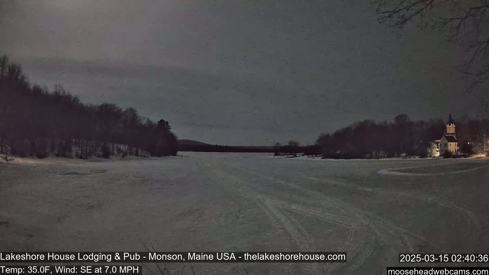 A snow-covered frozen lake is seen at night under a dark, overcast sky, with trees and a building visible in the distance.