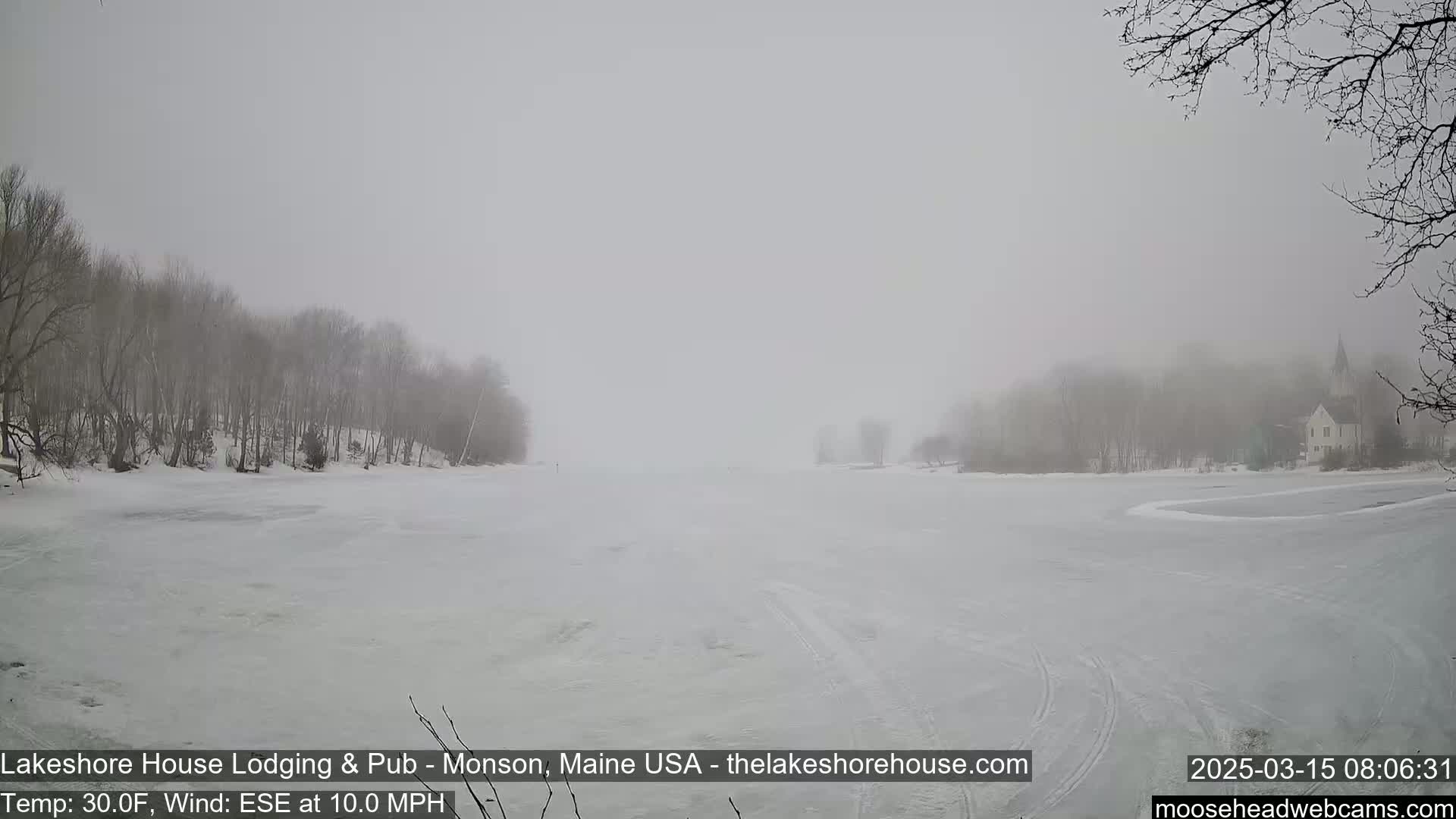 A snow-covered, frozen lake is visible under a foggy sky, with snow-covered trees lining the shores and a building in the distance.
