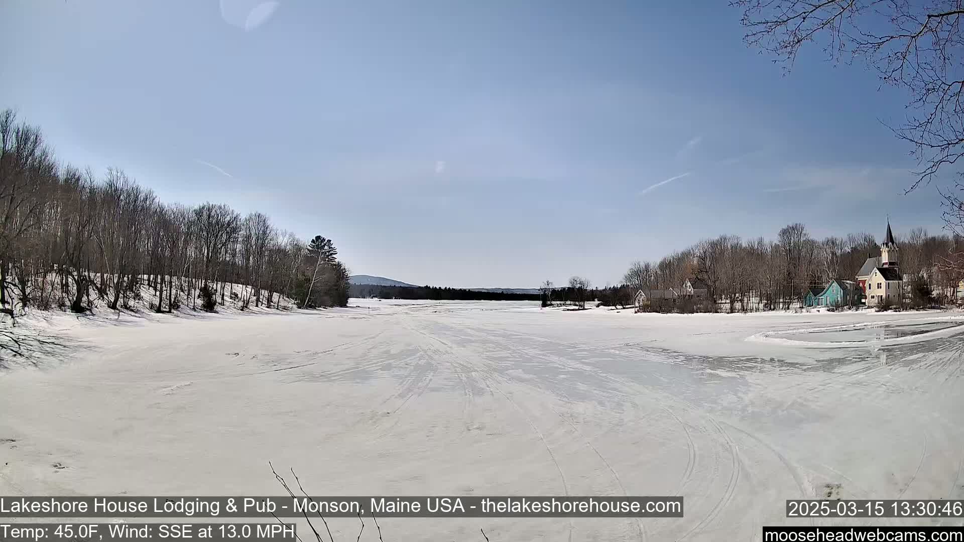 A frozen lake stretches out under a clear blue sky, with snow-covered trees lining the shore and a few buildings visible in the distance.