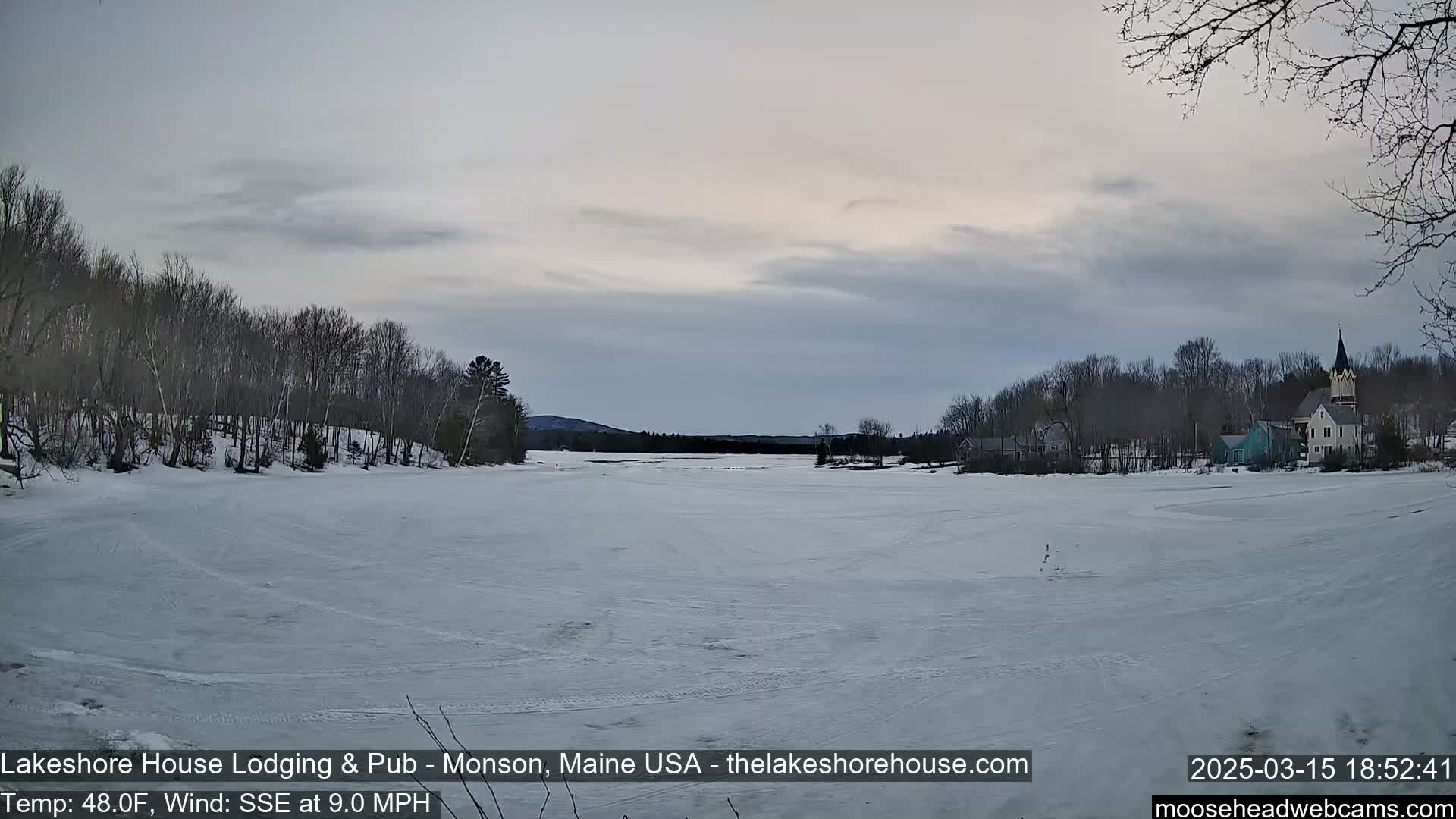 A snow-covered lake stretches towards a village of houses and trees under a cloudy sky.