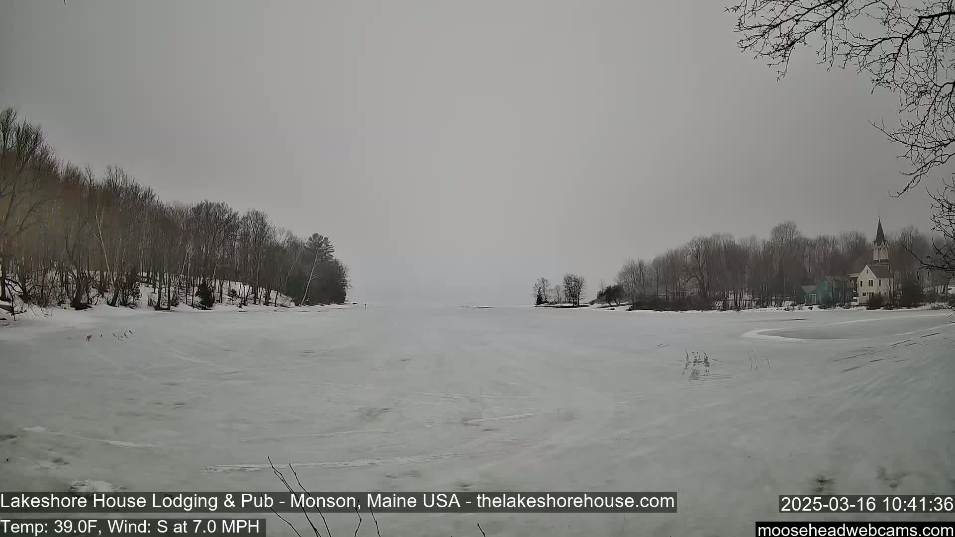 A mostly frozen lake is seen under an overcast sky, with snow-covered trees and buildings in the background, and a slight wind blowing from the south.