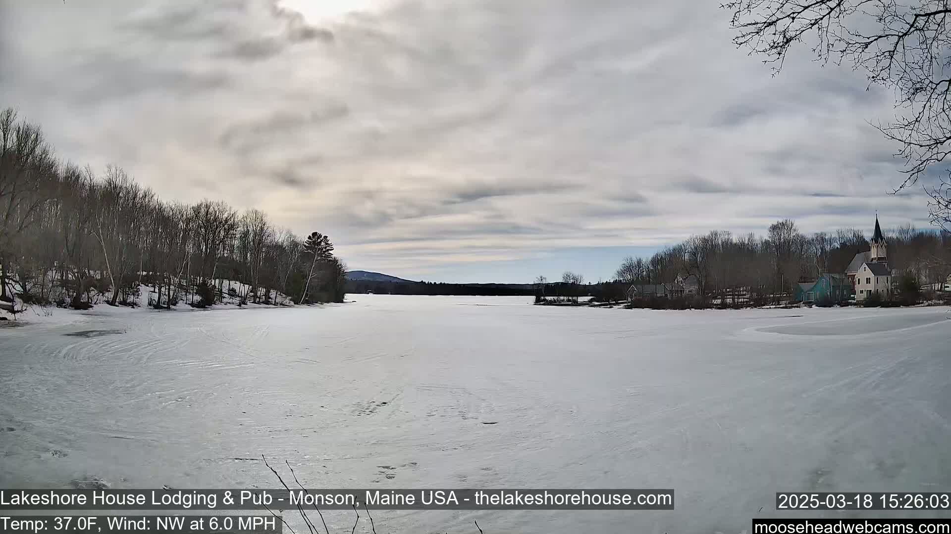 A frozen lake stretches out before a shoreline of bare trees and a small village under a cloudy sky.