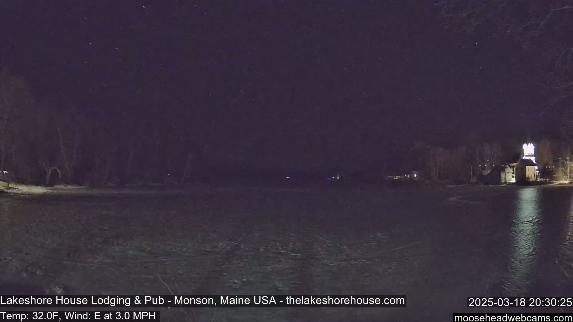 A nighttime view of a dark body of water under a starry sky, with a building lit up on the far shore and a slight east wind.