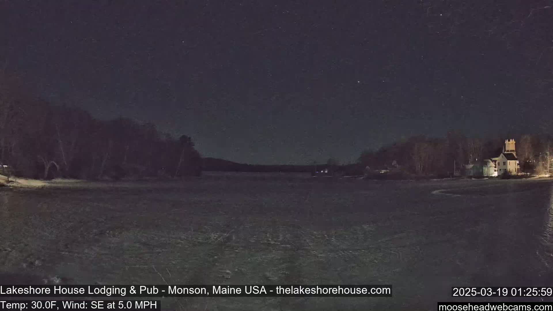 A nighttime view of a partly frozen lake with a building and trees on the far shore under a clear, starry sky, with a southeast wind at 5 mph and a temperature of 30 degrees Fahrenheit.