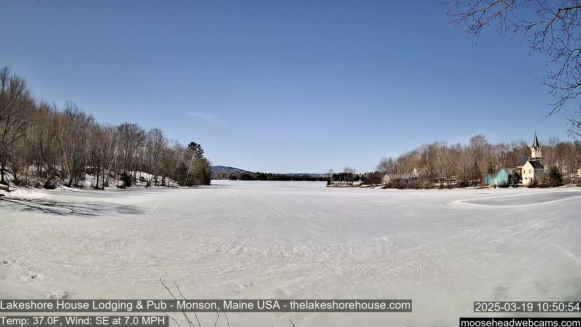 A frozen lake stretches out under a clear blue sky, bordered by bare trees and a small village with buildings including a church steeple in the background.