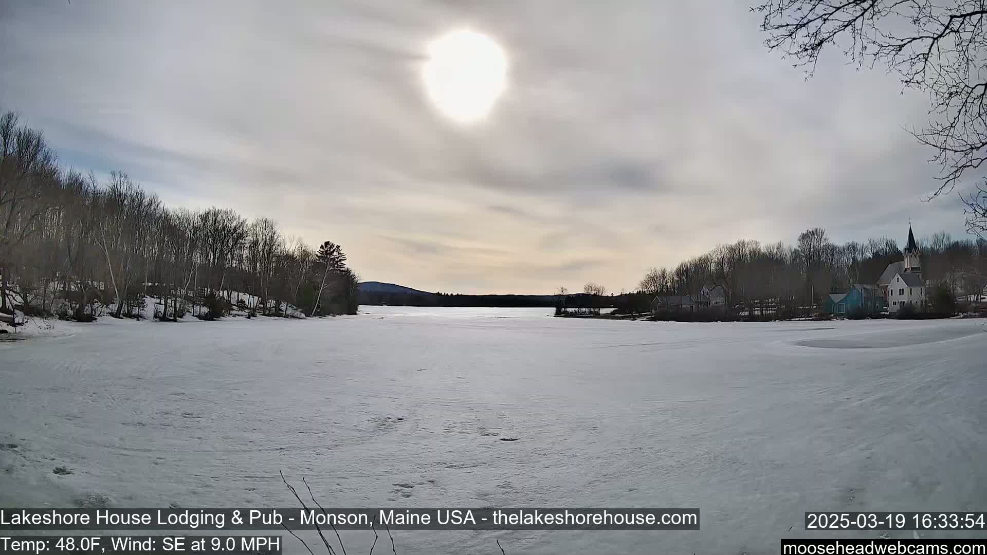 A mostly sunny day reveals a frozen lake with a snow-covered shoreline, trees, and a village in the background.