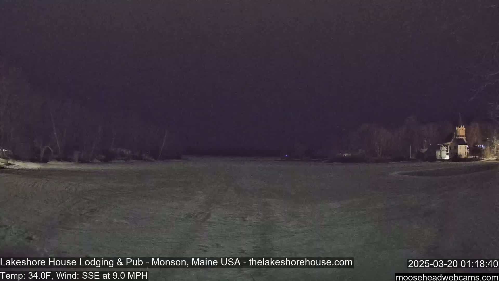 A snow-covered field at night, with a building illuminated in the distance under a dark sky.