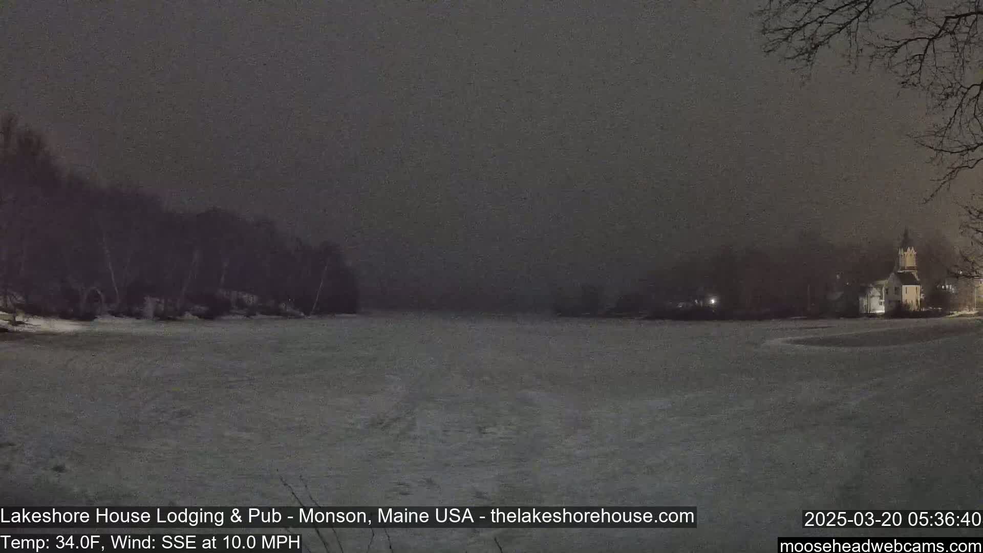 A snow-covered, dark landscape at night shows a body of water and a building in the distance under a cloudy sky with a 10 mph wind.