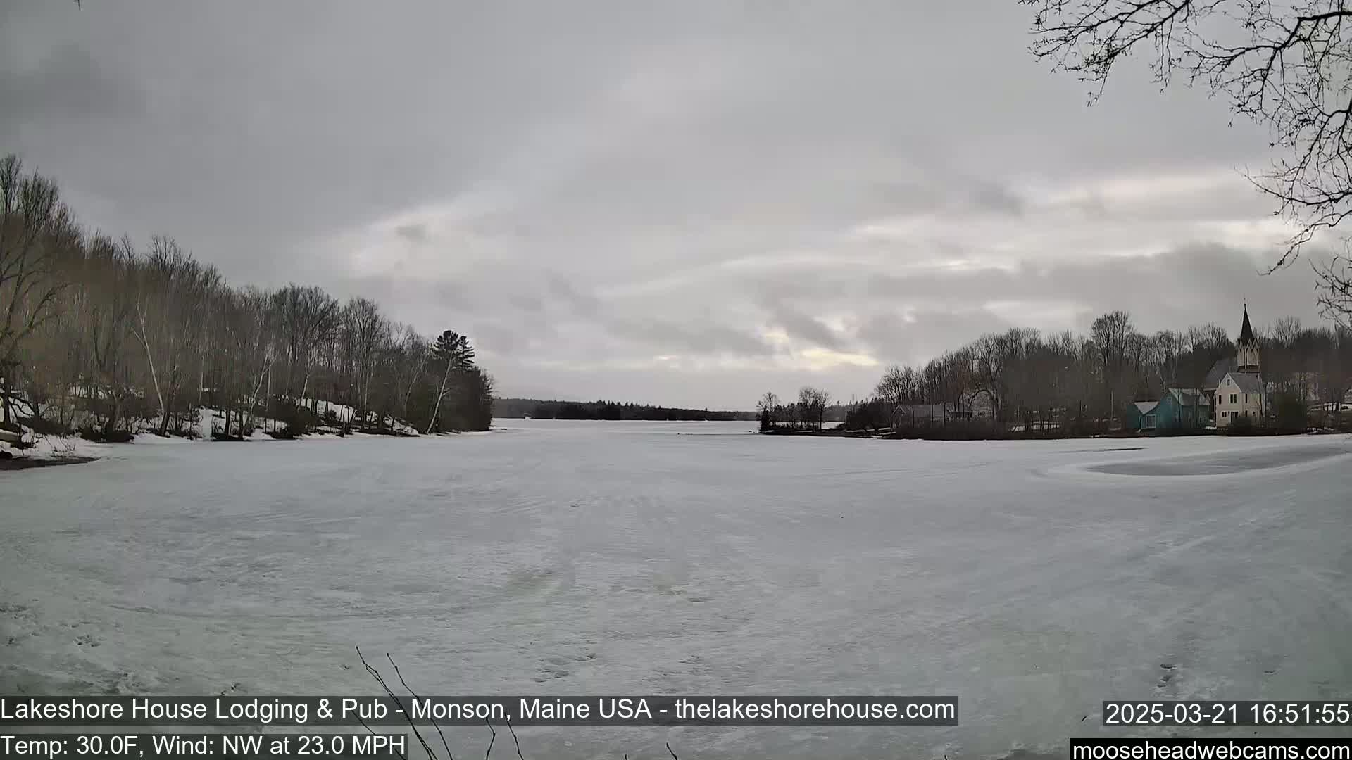 A frozen lake stretches out under a cloudy sky, with bare trees lining the shoreline and a village visible in the distance, including a church steeple.