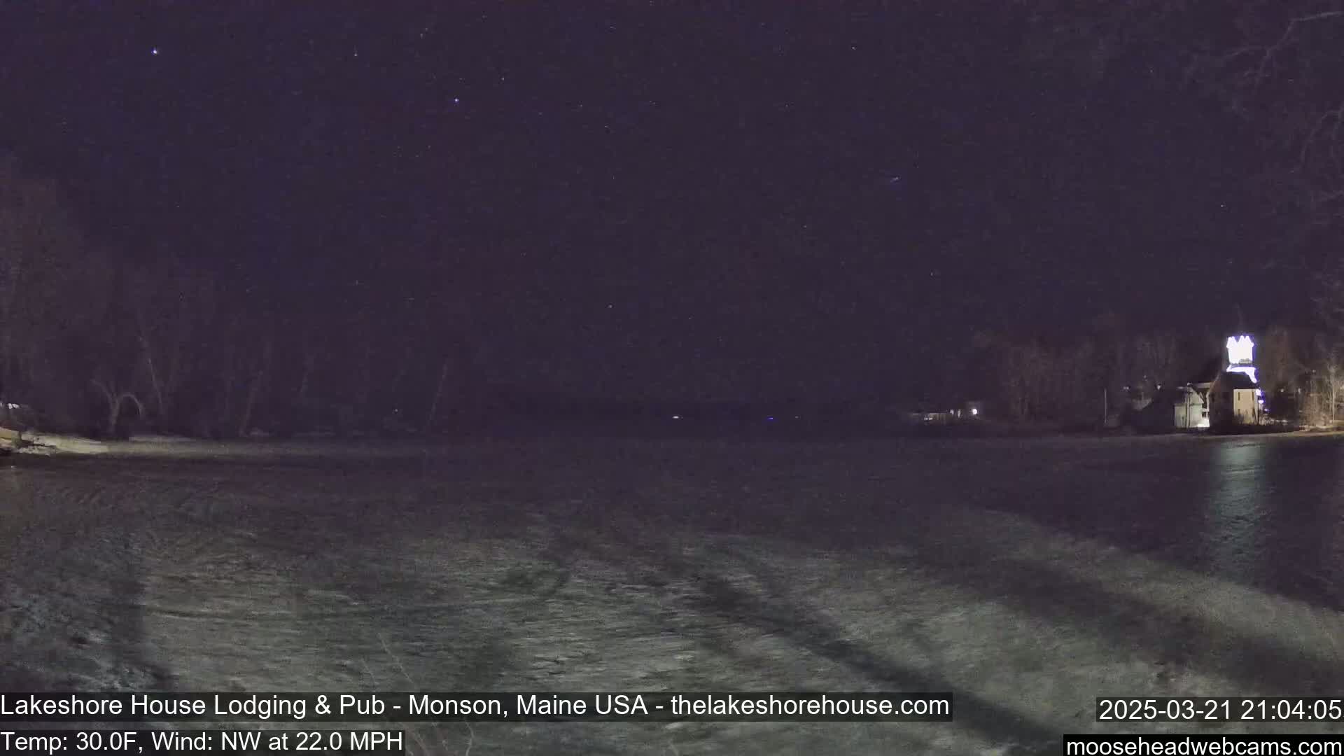 A nighttime view of a frozen lake with a building lit up on the far shore under a clear, starry sky, with a strong northwest wind.