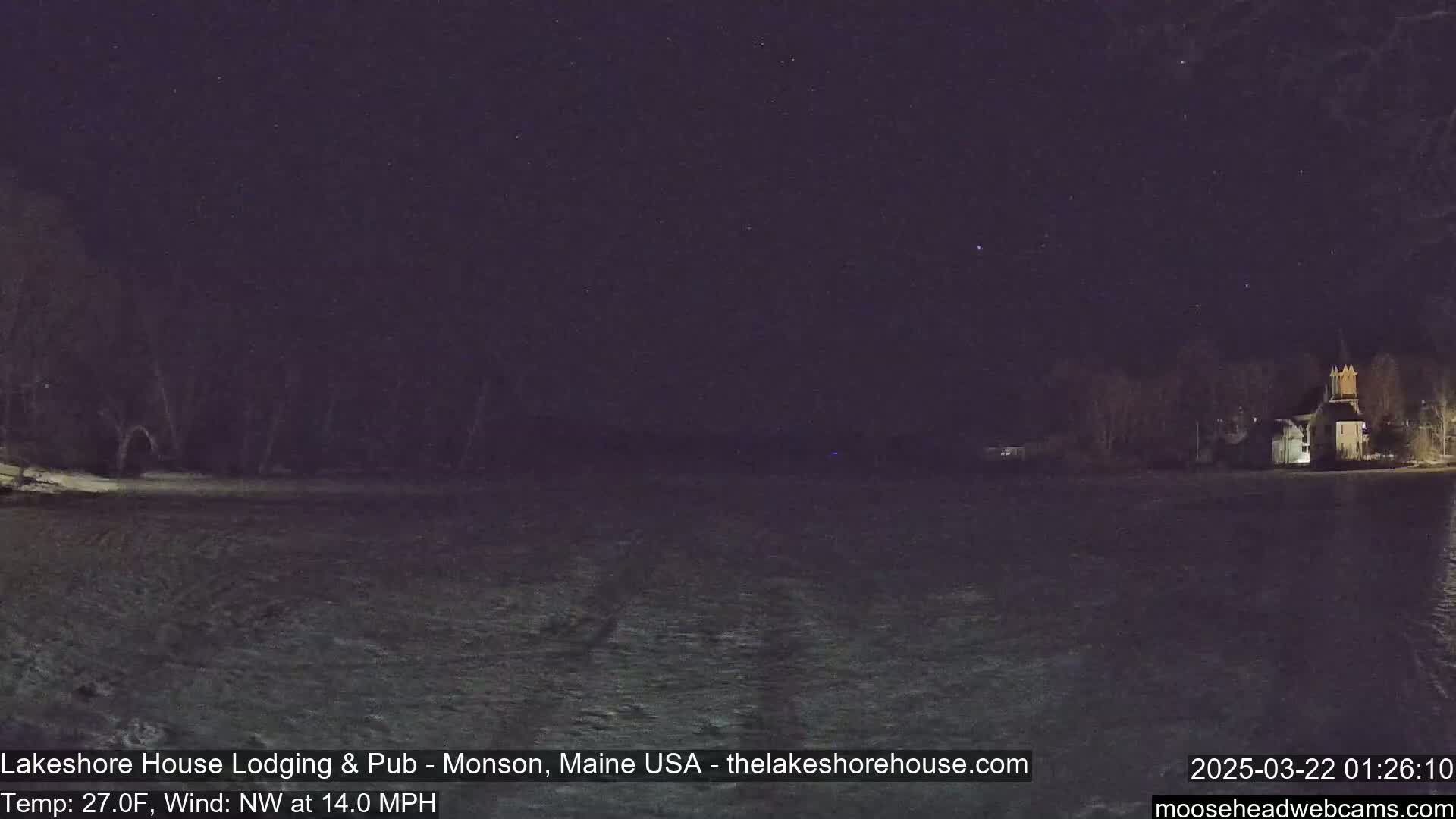 A nighttime, snow-covered landscape shows a building with lights on in the distance under a starry sky with a northwest wind at 14 mph and a temperature of 27°F.