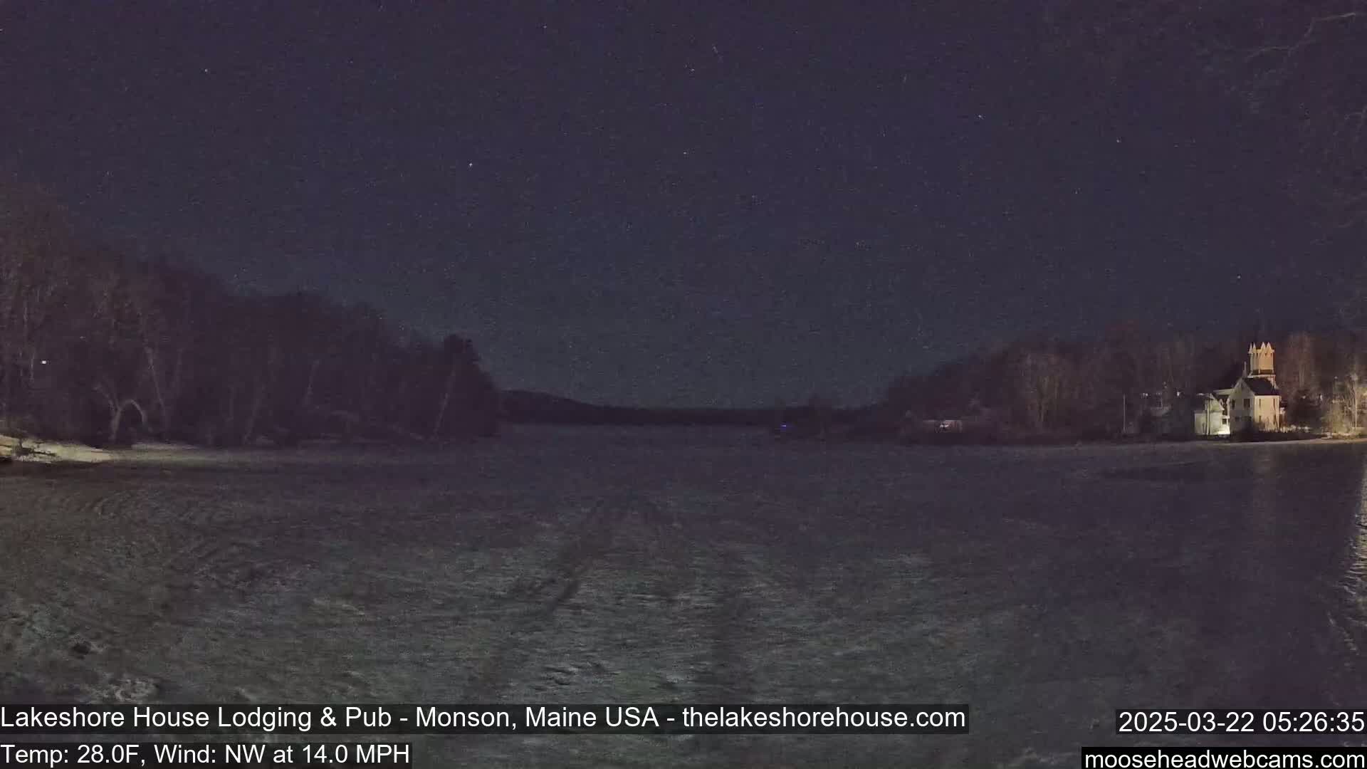A nighttime view of a frozen lake under a starry sky, with trees and a building visible on the far shore, and a 28°F temperature with a 14 mph northwest wind.