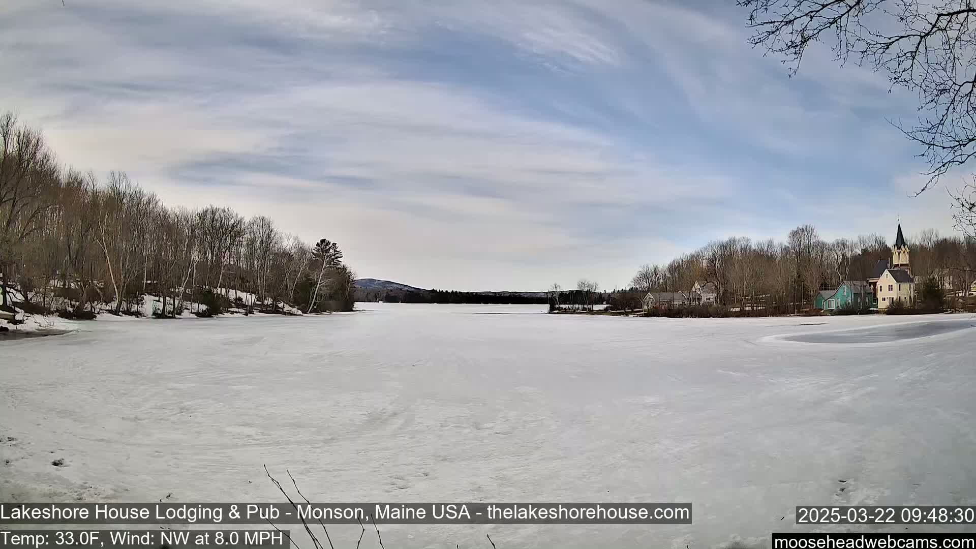 A frozen lake stretches out before a shoreline of bare trees and a small village under a partly cloudy sky.