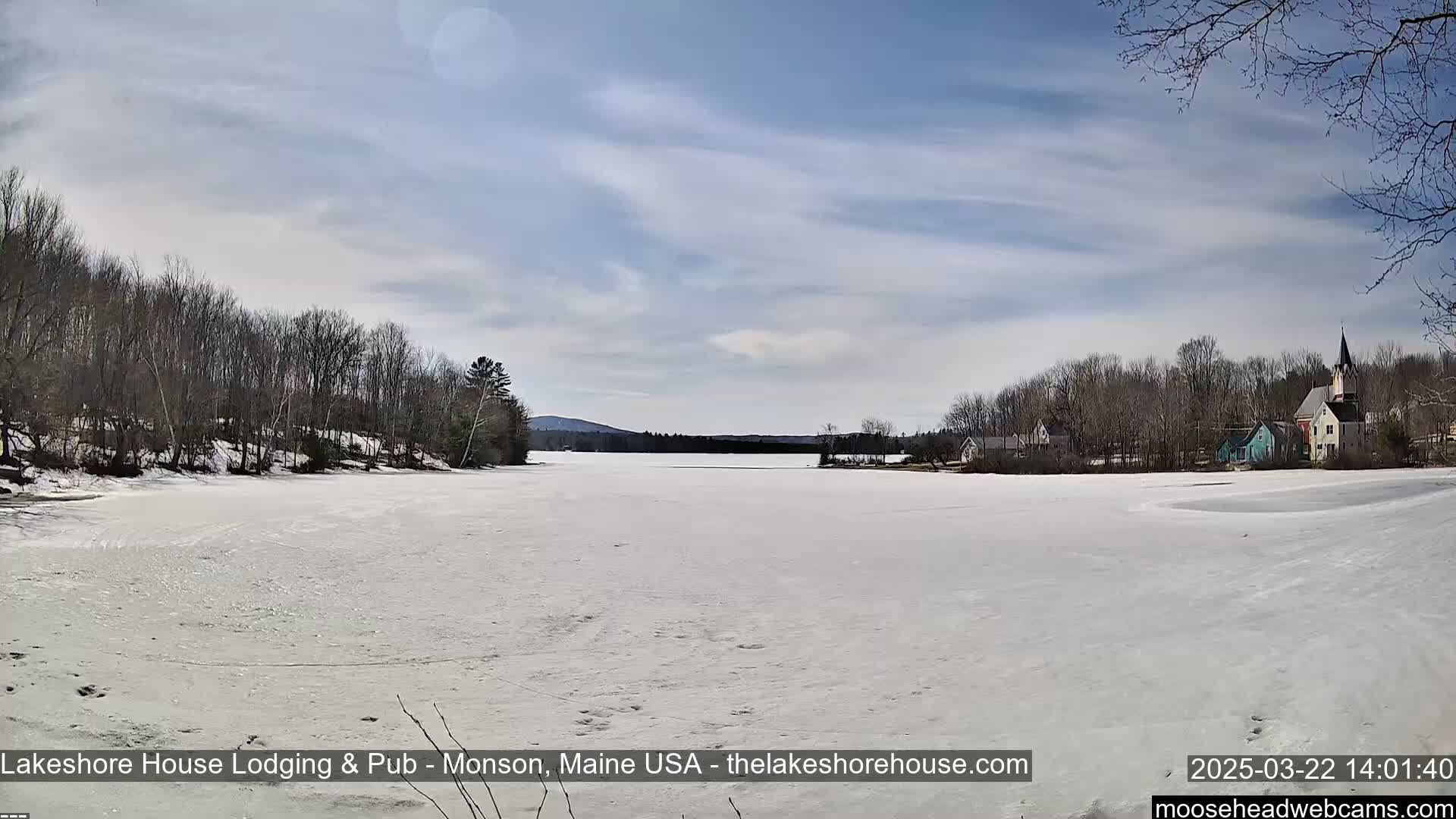 A frozen lake stretches across a snowy landscape under a mostly clear, bright sky, with leafless trees lining the shore and a small village visible in the distance.