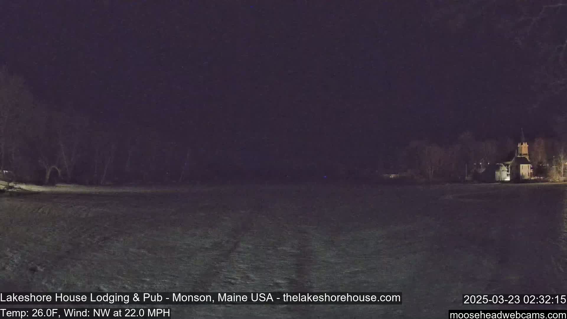 A snow-covered field at night is visible under a dark sky, with a building illuminated in the distance and trees silhouetted against the darkness; a northwest wind is blowing at 22 mph with a temperature of 26 degrees Fahrenheit.