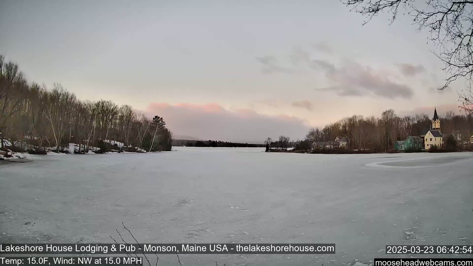 A frozen lake with a town and bare trees on the far shore is visible under a pale pink and gray sky, with a northwest wind blowing at 15 mph and a temperature of 15°F.