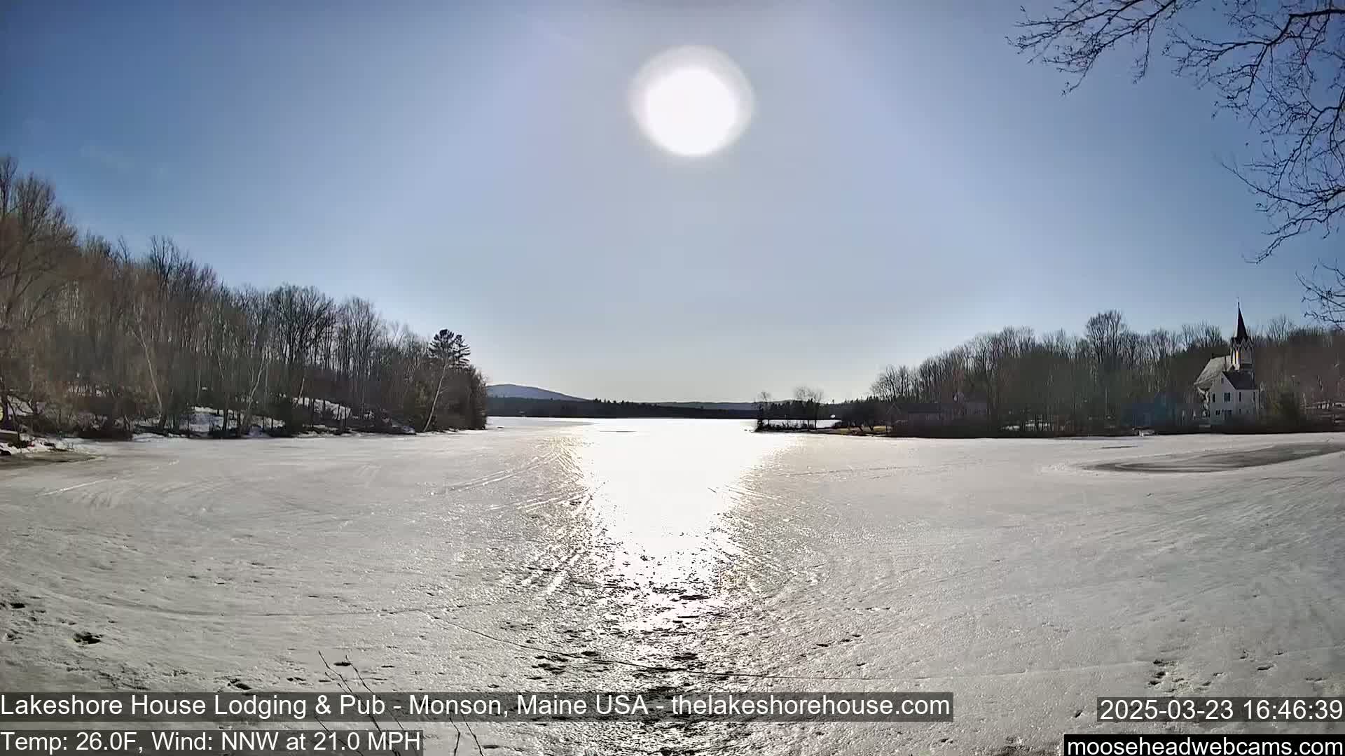 A frozen lake reflects the bright sun under a clear sky, with snow-covered banks lined with leafless trees and a building visible in the distance.