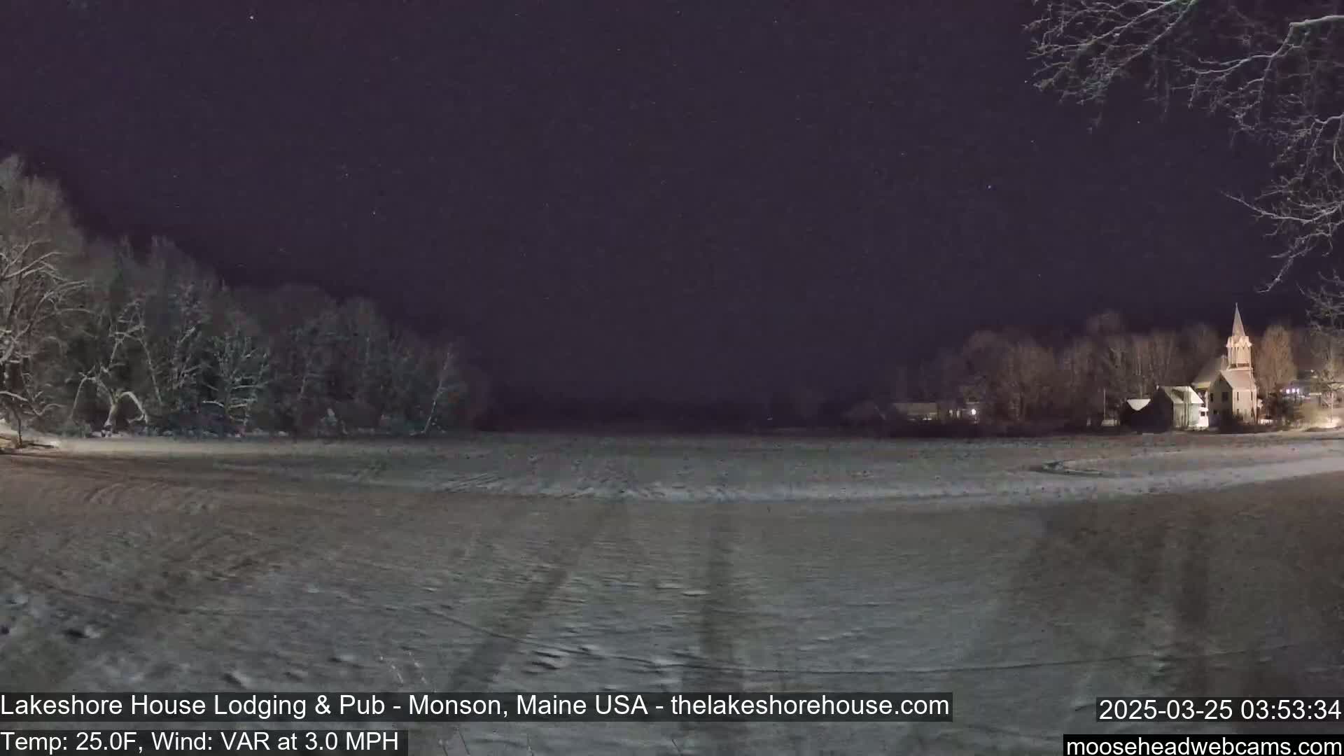 A snow-covered field at night under a starry sky, with a line of snow-covered trees to the left and a church steeple visible in the distance to the right.