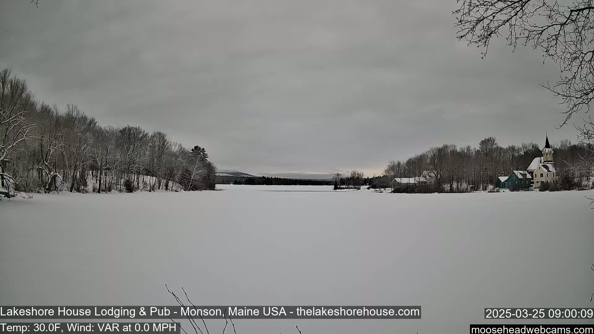 A snow-covered frozen lake stretches to the horizon, under a gray sky, with snow-covered trees lining the shore and buildings visible in the distance.