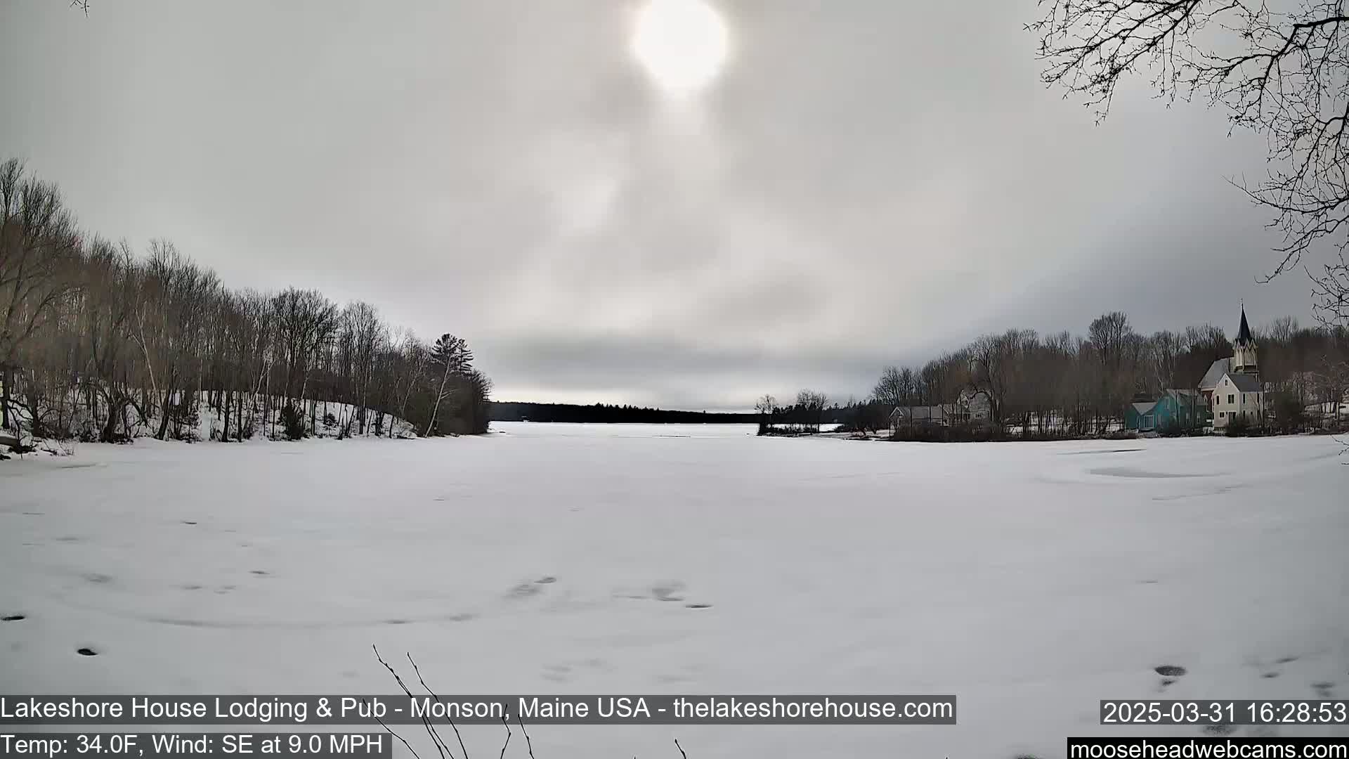 A frozen lake under a mostly cloudy sky is seen with a small town visible across the water and bare trees lining the shore.