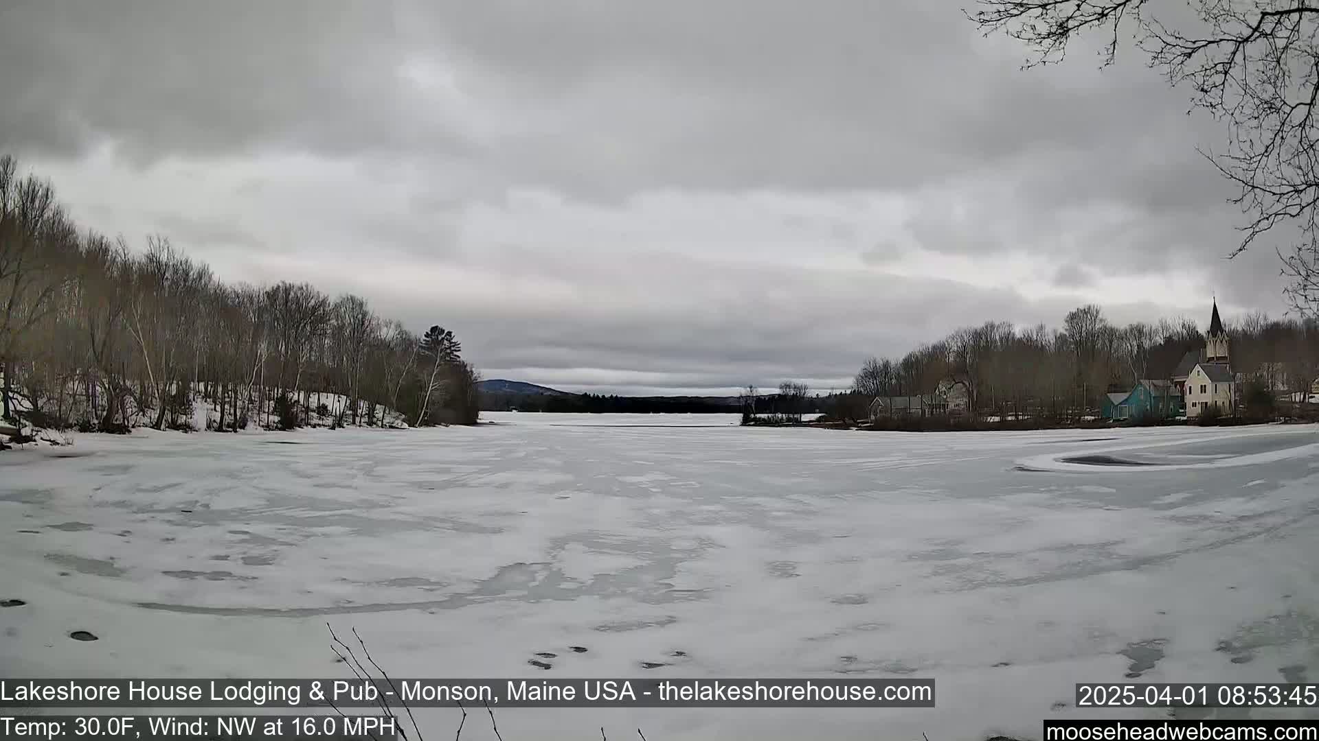 A mostly frozen lake is seen under a gray, overcast sky, with bare trees lining the shoreline and a small town visible in the distance.
