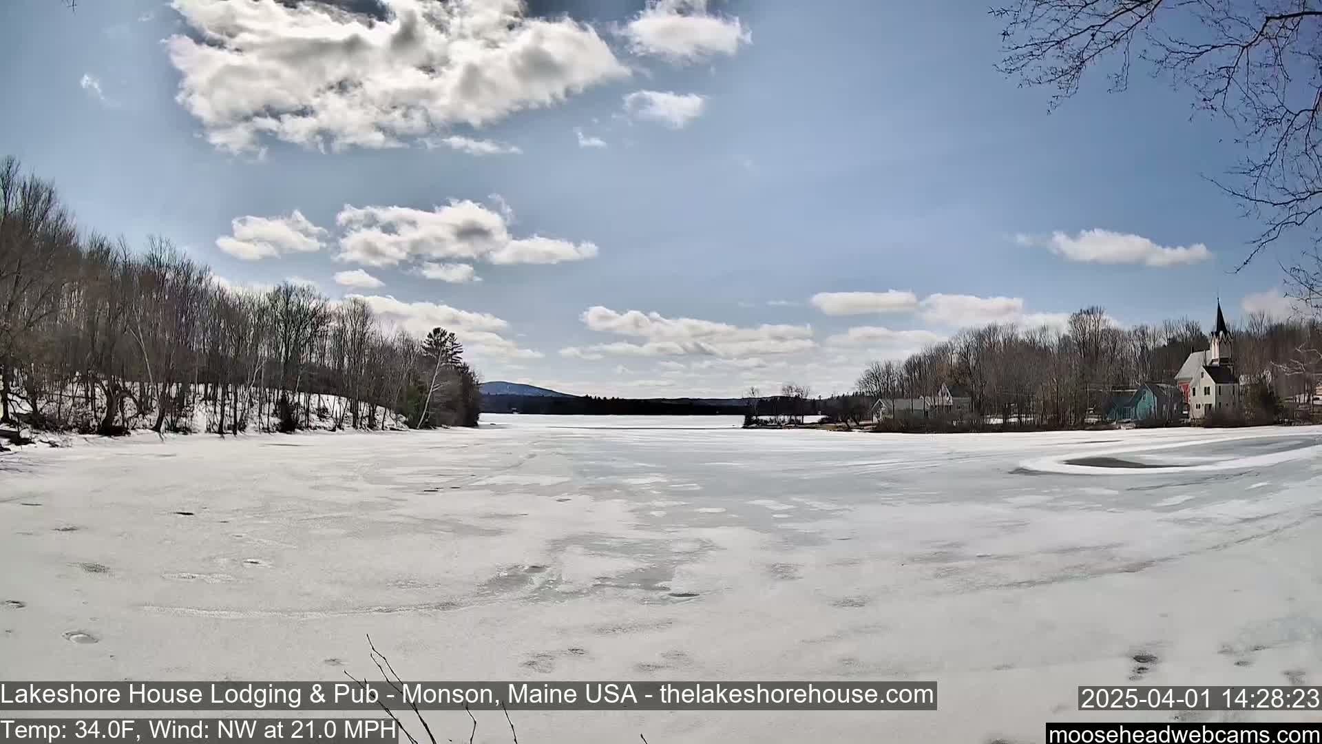 A mostly frozen lake is visible, under a partly cloudy sky, with bare trees and buildings lining the far shore.