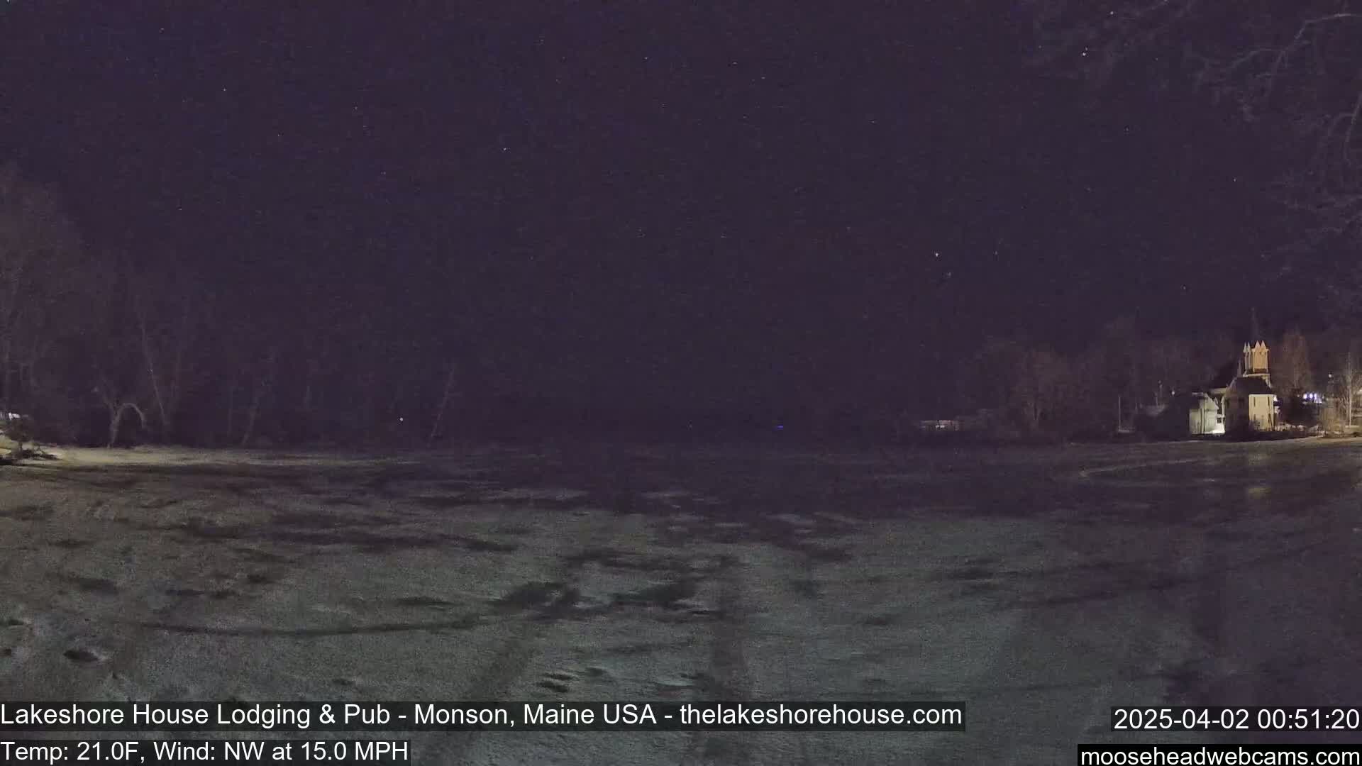 A snow-covered field at night under a starry sky, with a building faintly lit in the distance, and a northwest wind blowing at 15 mph.
