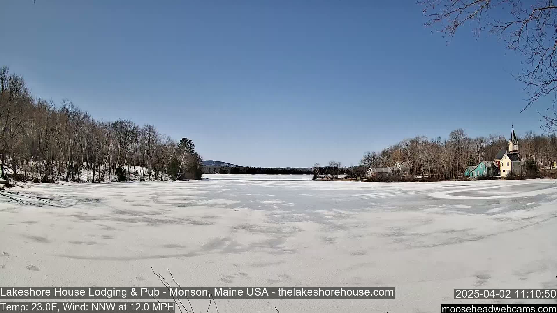 A frozen lake, mostly covered in snow, stretches out to a small town nestled amongst bare trees under a clear blue sky.
