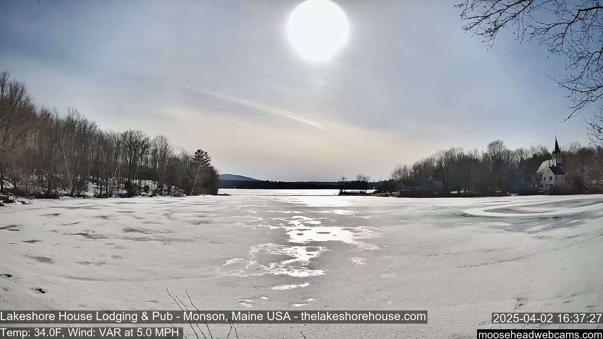 A partially frozen lake is seen under a bright, sunny sky, surrounded by bare trees and a small town in the background.