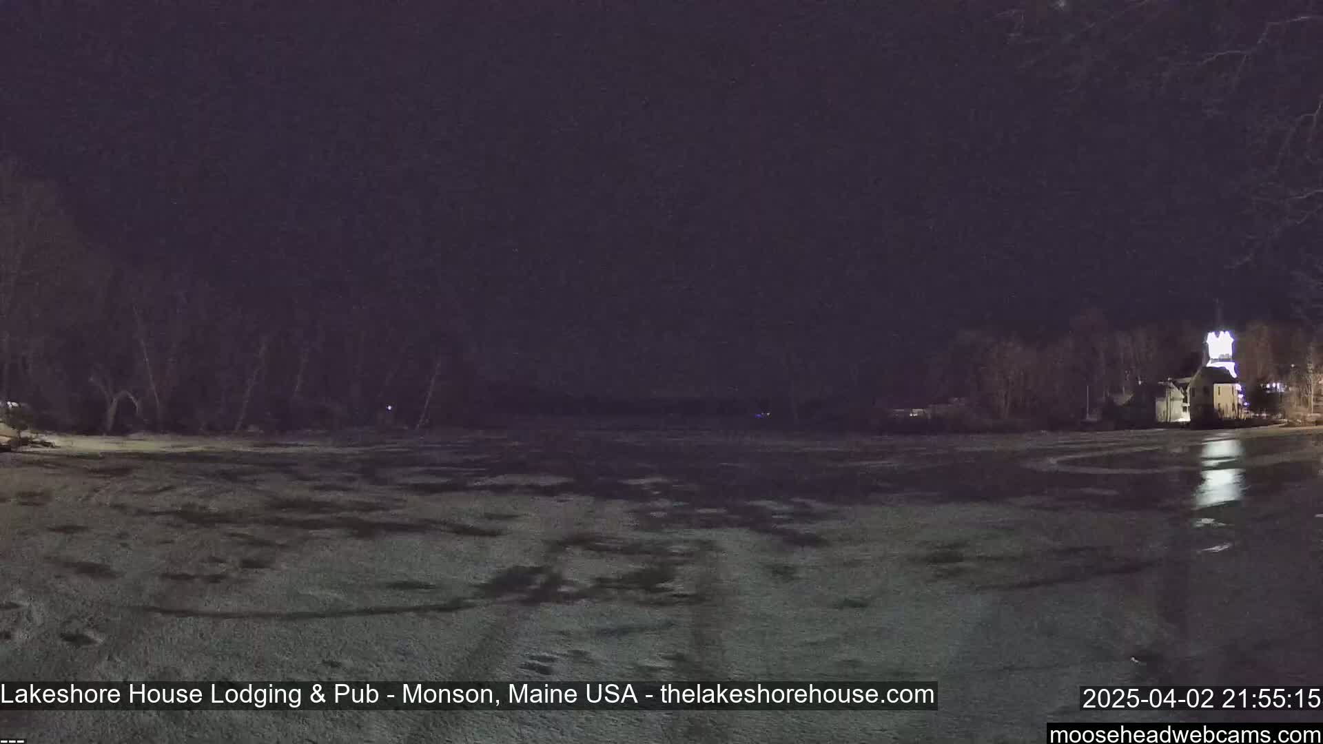 A snow-covered, partially frozen lake is seen at night, with a building lit up in the distance under a dark sky.