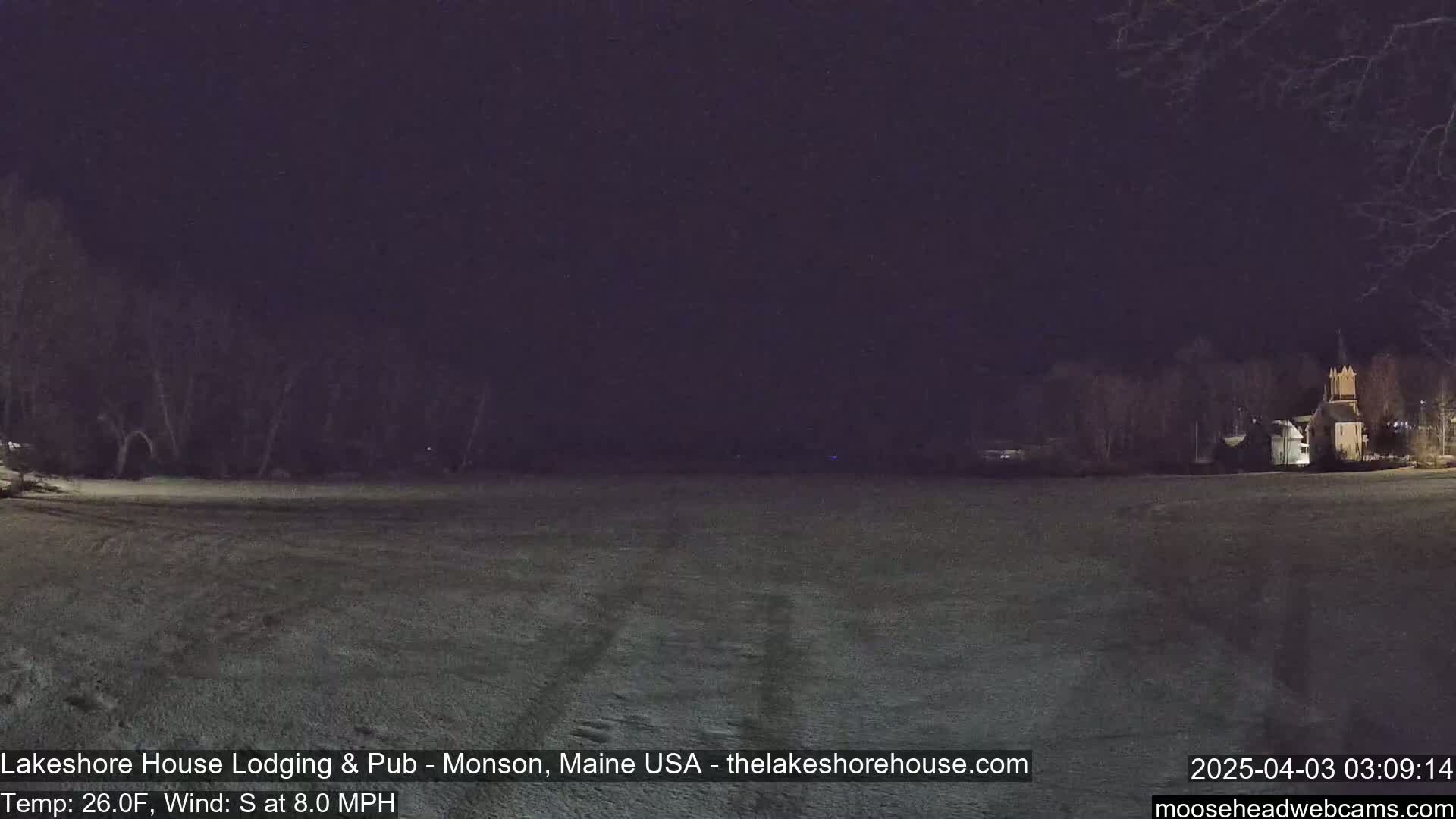 A snow-covered field at night is illuminated, showing a sparsely wooded area and a building in the distance under a dark sky.