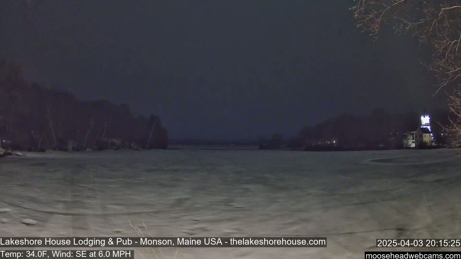 A dark, nighttime view of a frozen lake, with a building lit up on the far shore, under a clear, dark sky and a 6 mph southeast wind.
