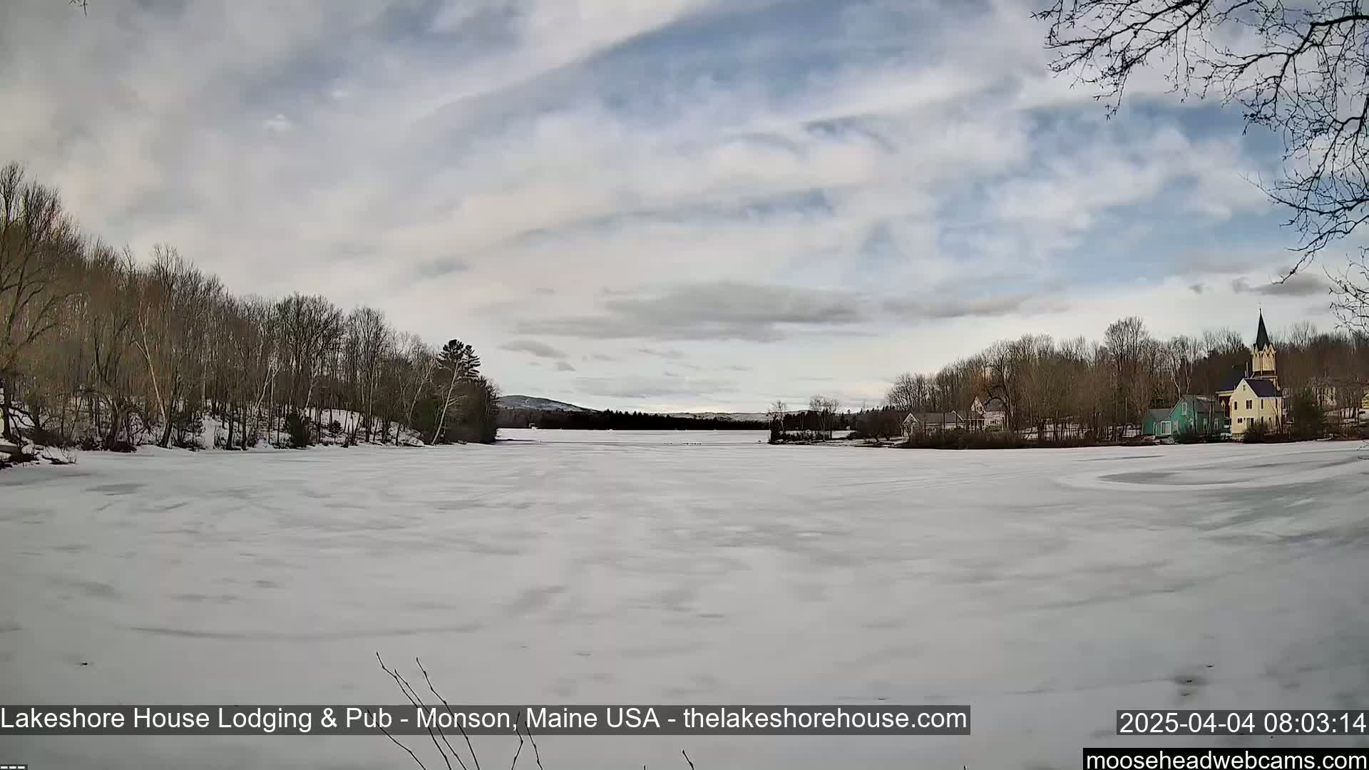 A frozen lake stretches out before a village of buildings under a mostly cloudy sky.