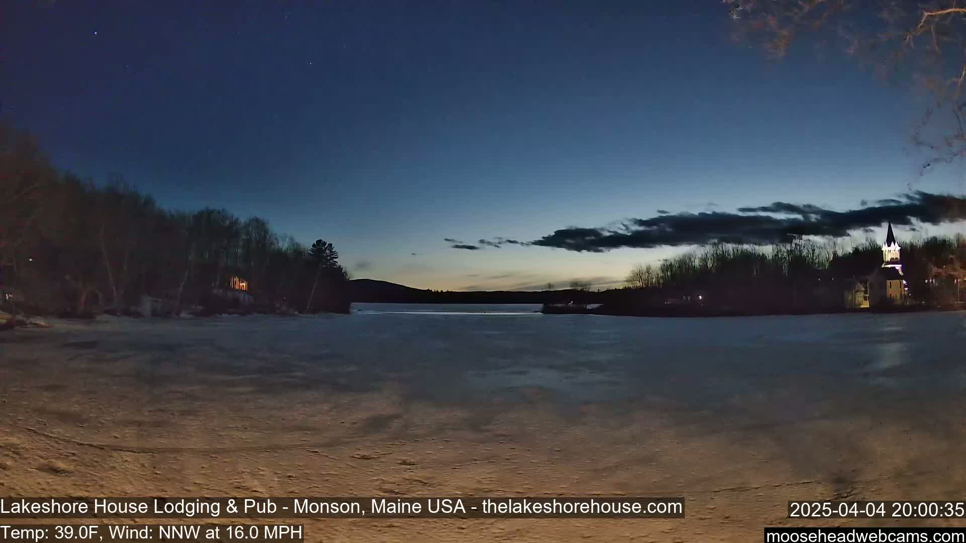 A partly frozen lake is visible at dusk under a mostly clear, dark blue sky with some clouds, with trees and a building lit up on the far shore.