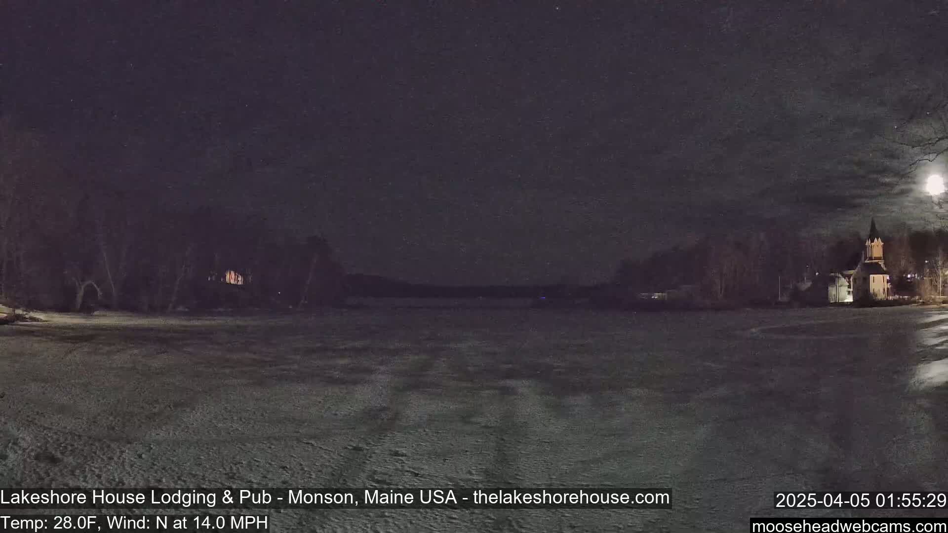 A snow-covered, frozen lake is seen at night under a mostly cloudy sky, with buildings and trees visible on the far shore.