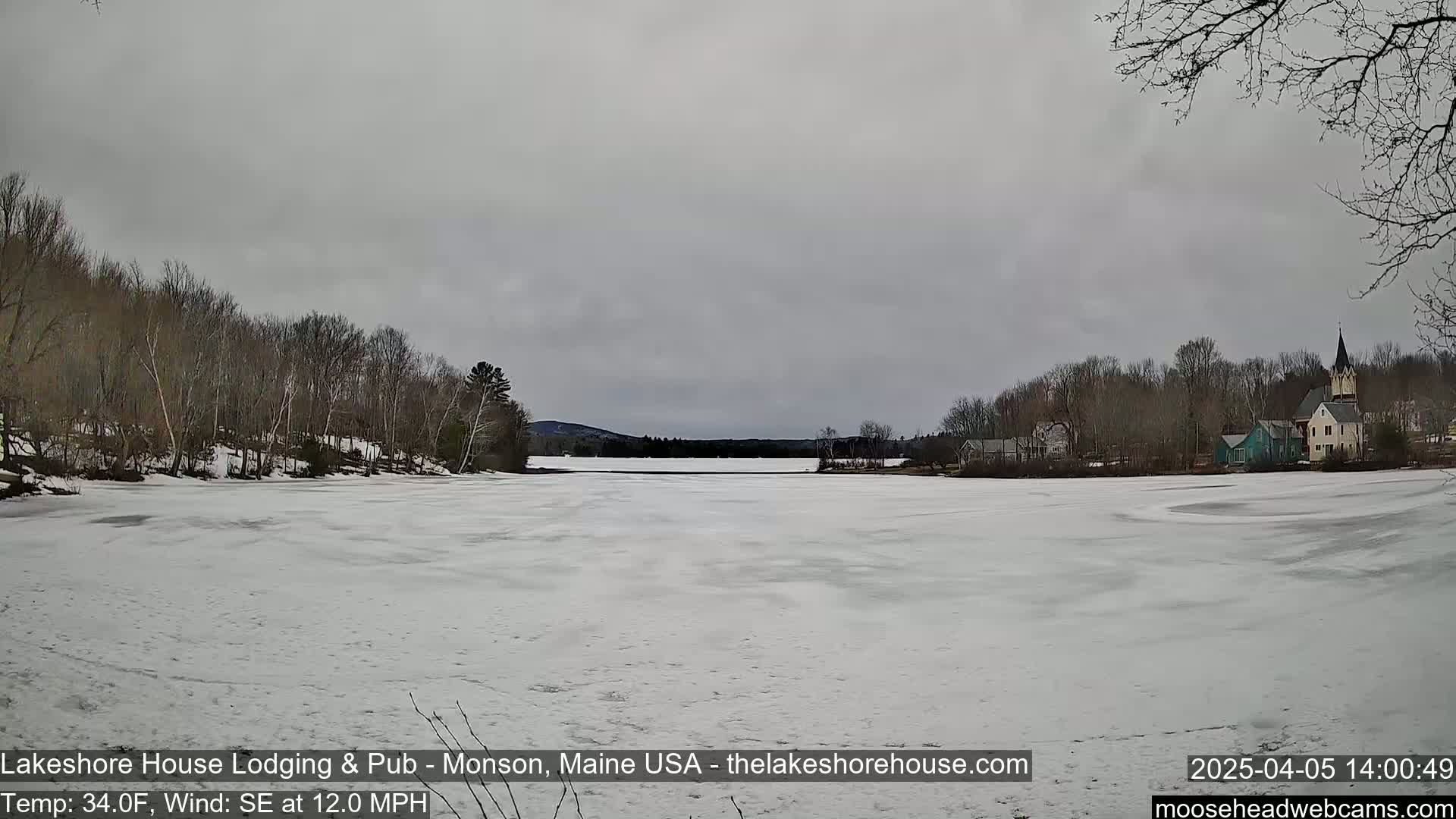 A frozen lake, covered in snow and ice, stretches out before a village with houses and a church steeple under an overcast sky.