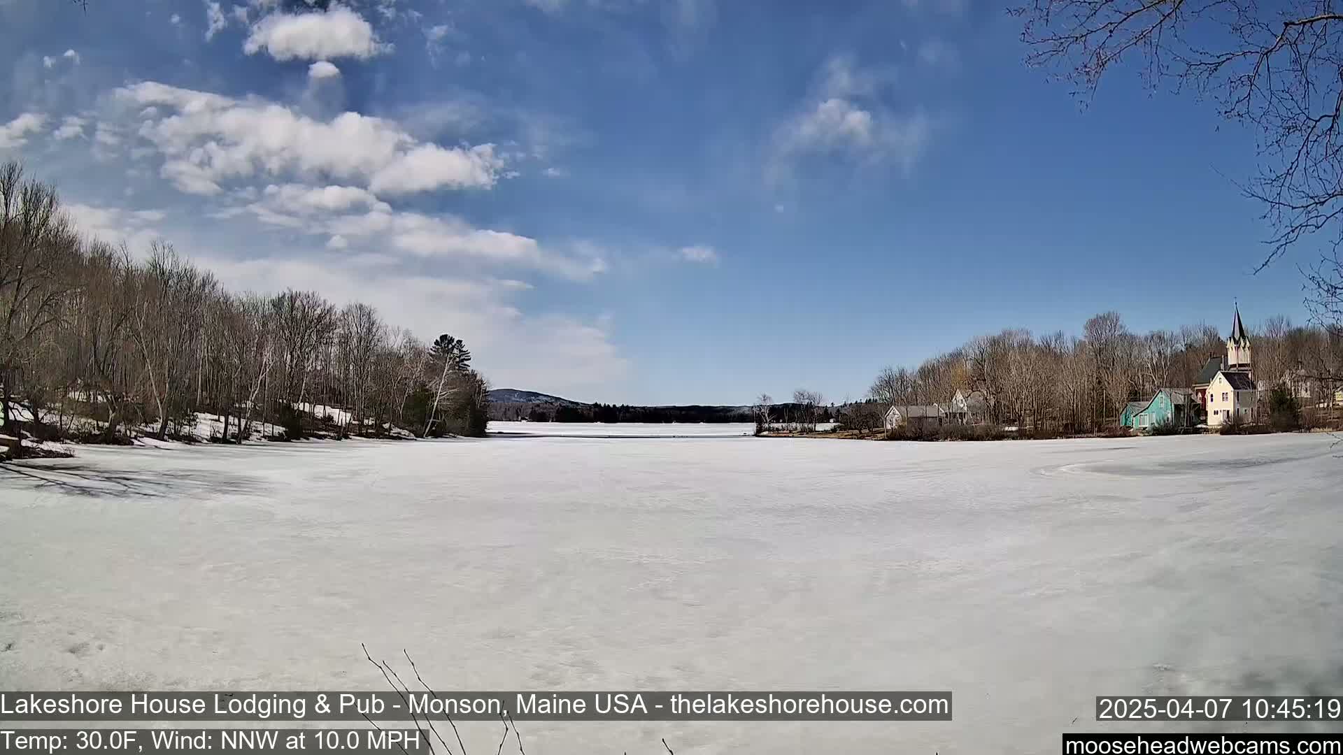 A mostly frozen lake stretches out under a partly cloudy blue sky, with a small town and bare trees lining the far shore.