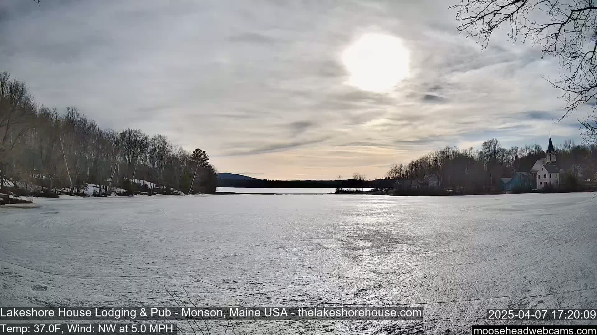 A partly sunny sky over a snow-covered frozen lake with bare trees lining the shores and buildings visible in the distance.