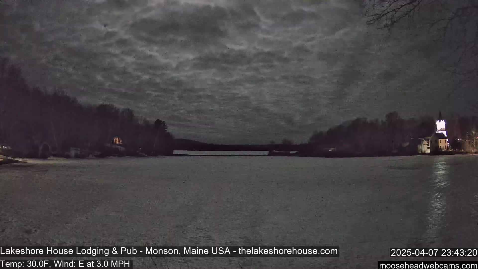 A frozen lake is visible under a dark, cloudy night sky, with snow-covered land and buildings with lights on in the distance.