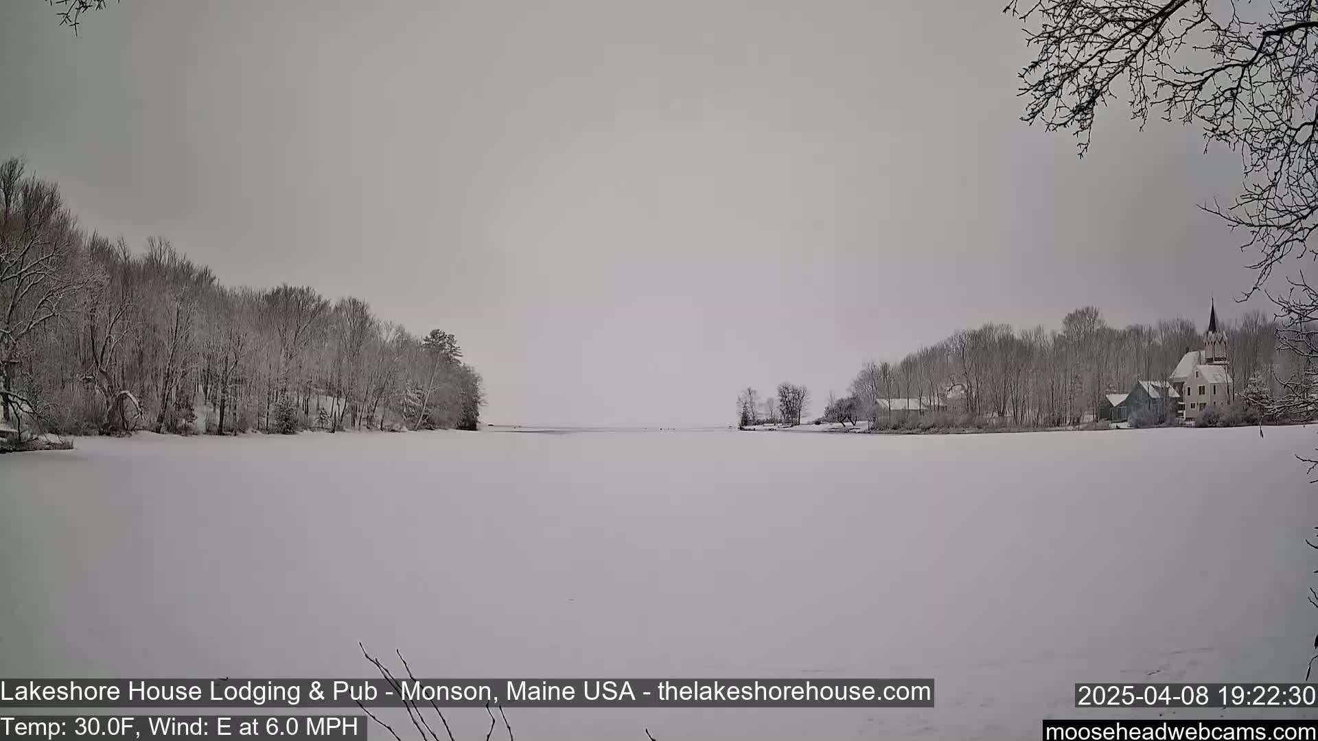 A snow-covered lake is seen with snow-covered trees and buildings on either side under an overcast sky.