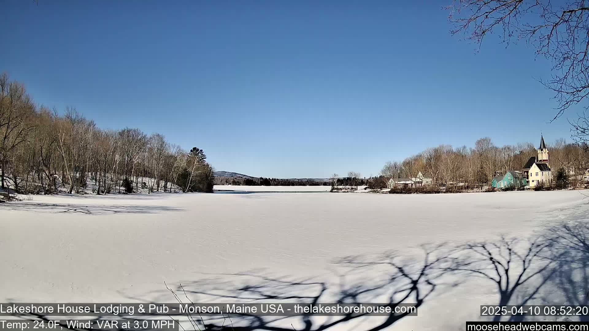 A frozen lake stretches out under a clear blue sky, with snow-covered trees and buildings visible on the far shore.