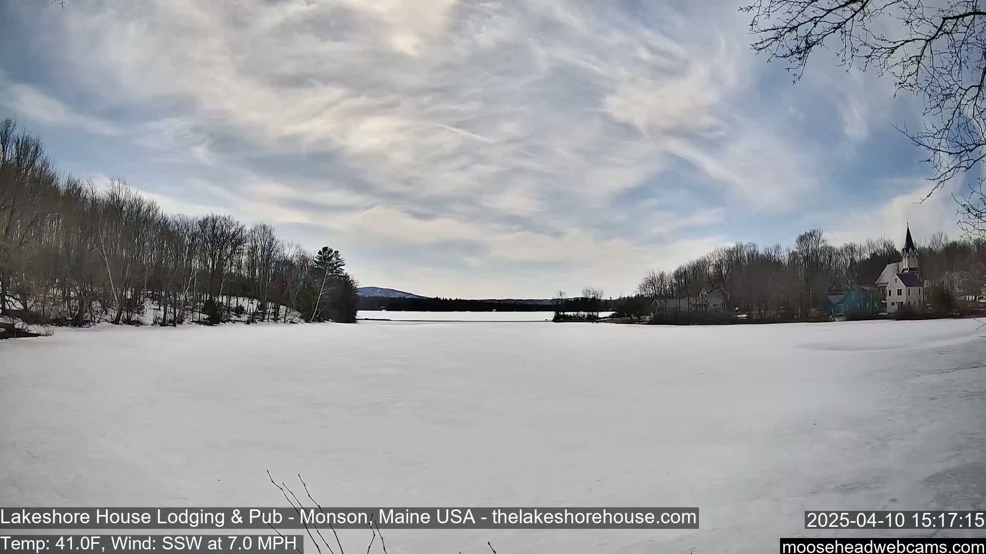 A frozen lake is seen under a partly cloudy sky, with bare trees lining the shore and buildings visible in the distance.