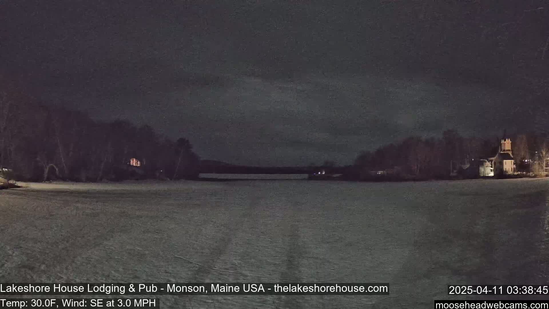A dark nighttime scene shows a snow-covered field, a body of water, and buildings with lights on across the water under a mostly clear, dark sky.