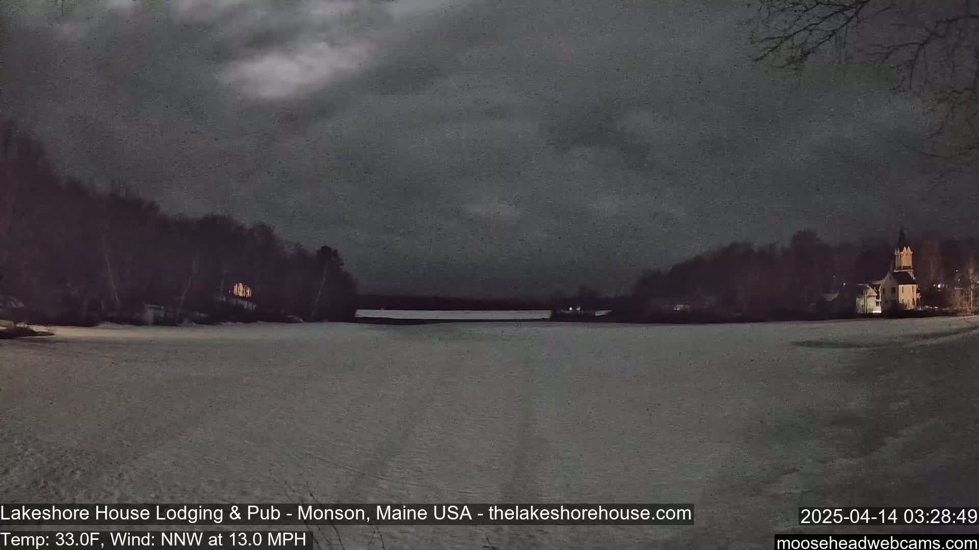 A snow-covered field stretches across a frozen lake, under a dark, cloudy night sky, with buildings and trees visible on both sides of the lake.