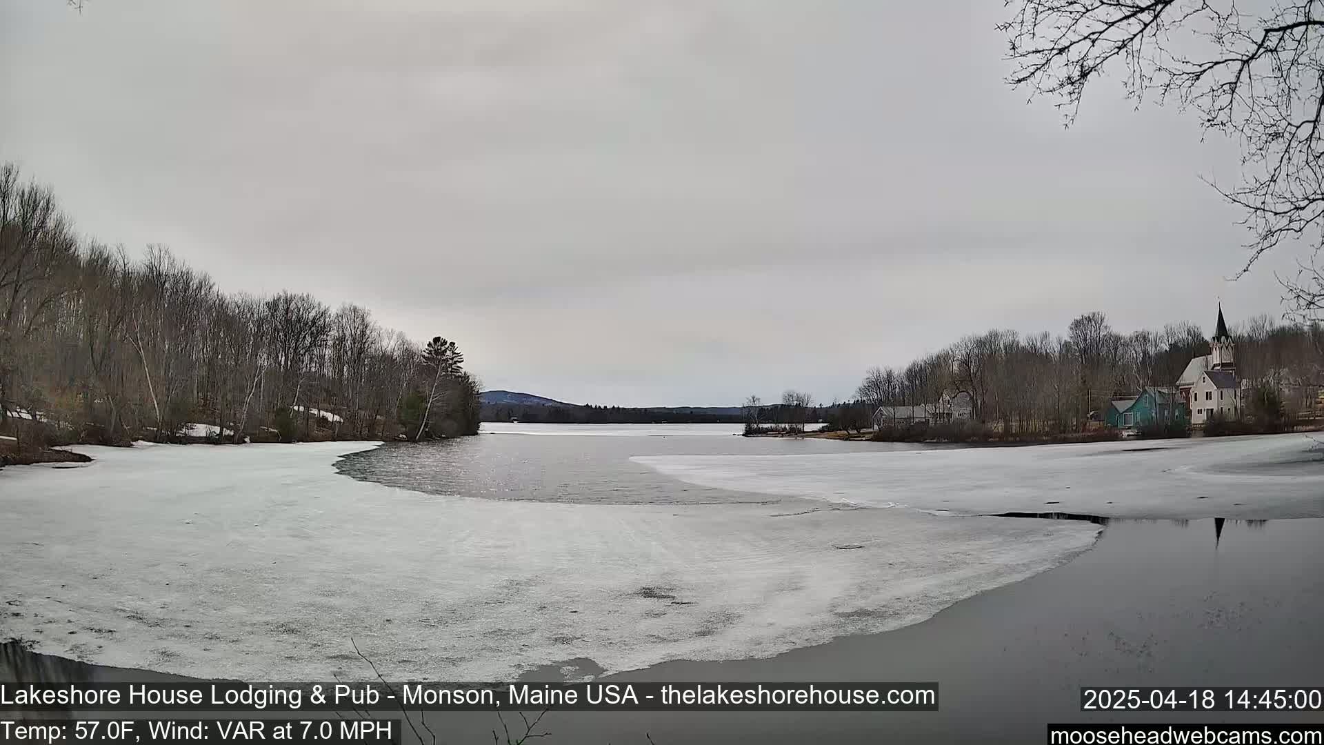 A partially frozen lake is visible, with bare trees lining the shore and a small village with a church steeple in the background under an overcast sky.