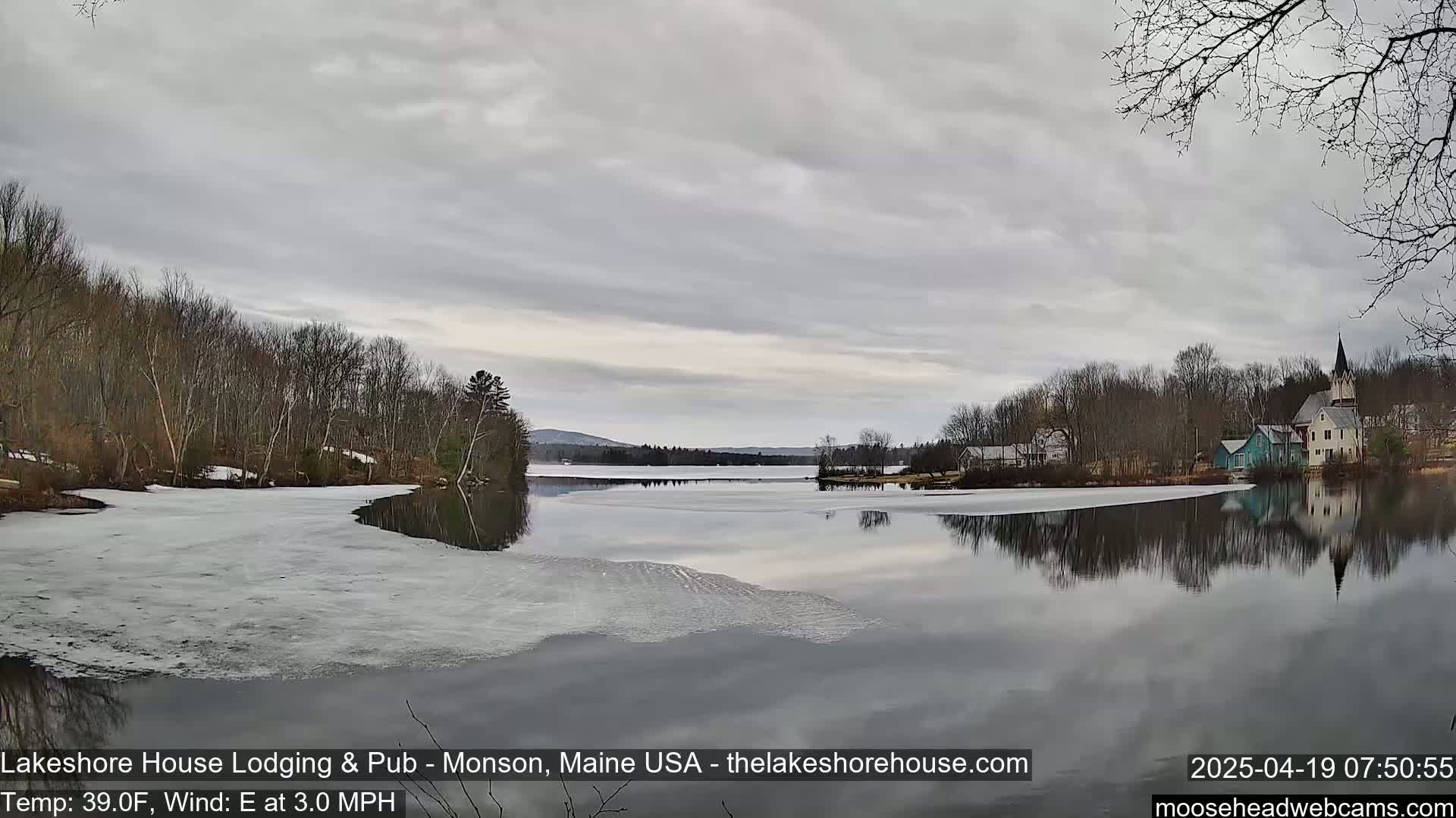 A calm lake with partially melted ice reflects a cloudy sky and buildings on its far shore, under a mostly cloudy sky.